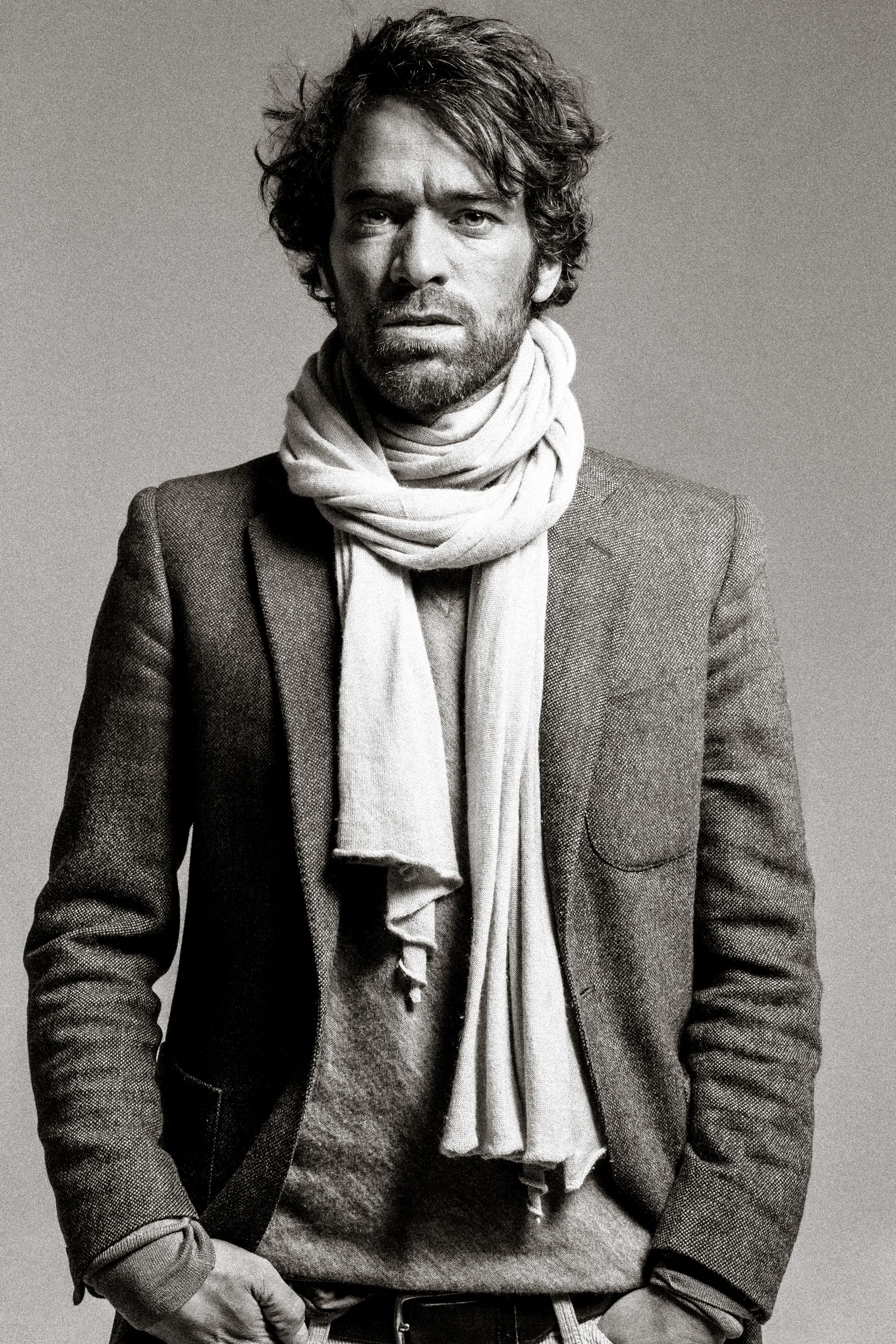 Black and white portrait of a person wearing a gray blazer and draped white scarf with tousled dark hair.