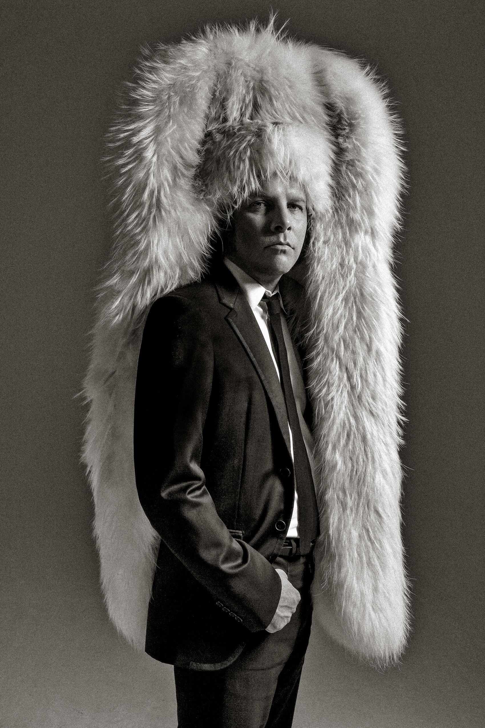 A person in a black suit stands against a dark background wearing an oversized white fur stole draped over their shoulders.