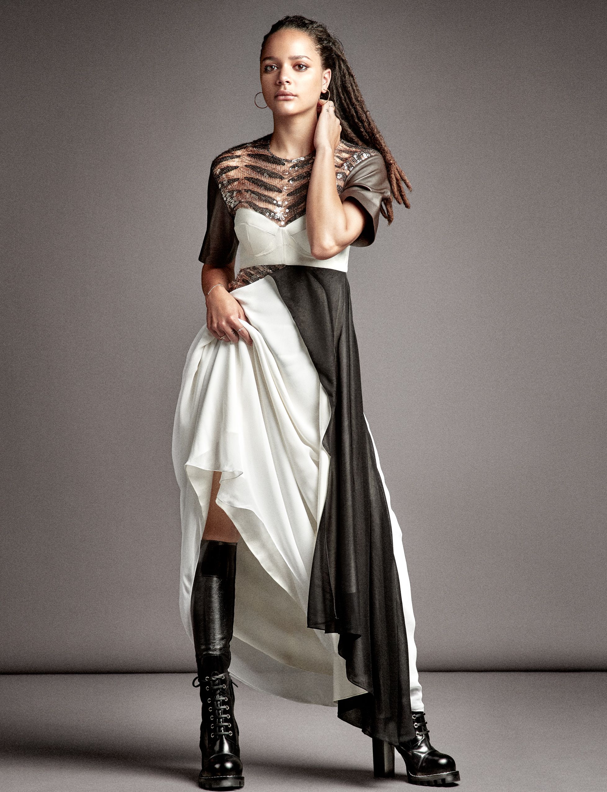 Fashion model in contrasting black and white dress with asymmetrical design poses in tall combat boots against gray backdrop.