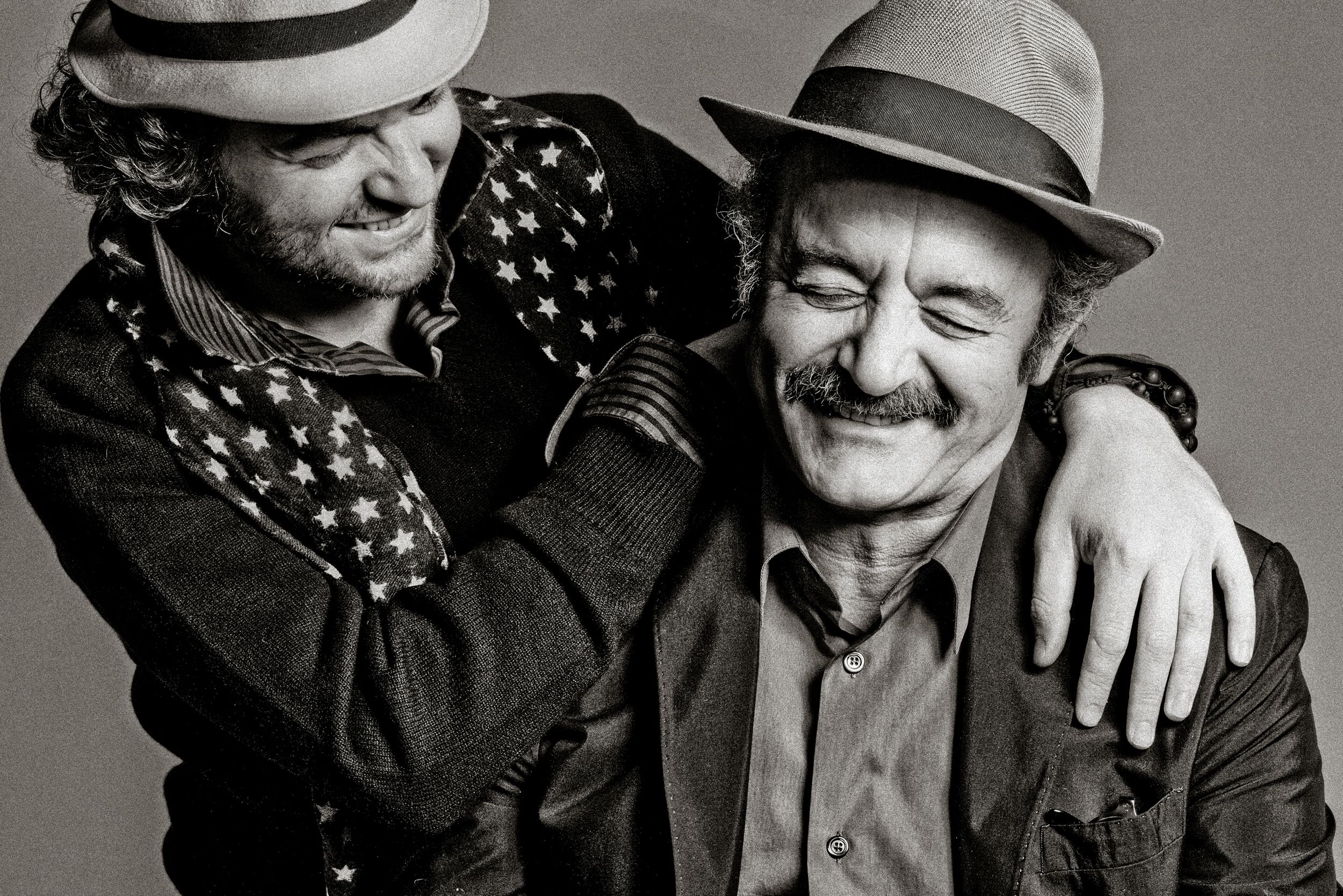 Two people wearing fedora hats share a warm embrace and smile in a black and white portrait.