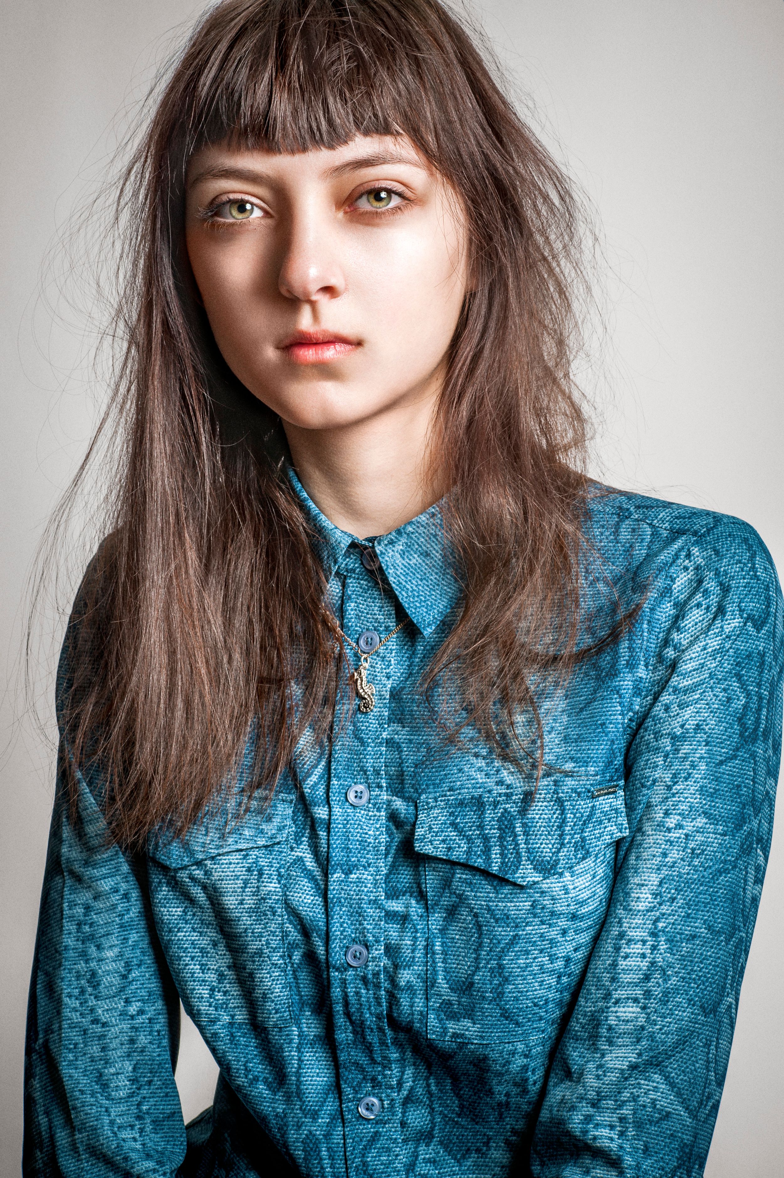 Young woman with long dark hair and bangs wearing a teal blue snakeskin print button-up blouse.