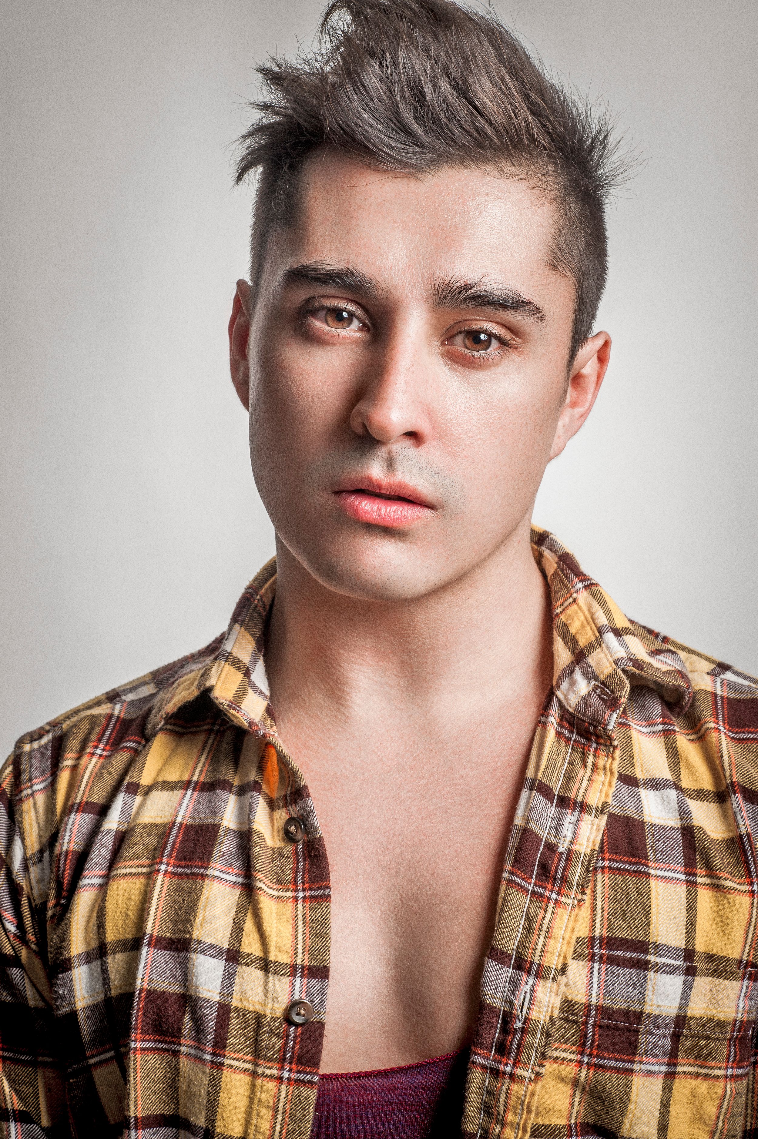 Young man with styled hair wearing an open plaid flannel shirt, posing for a portrait against a gray background.