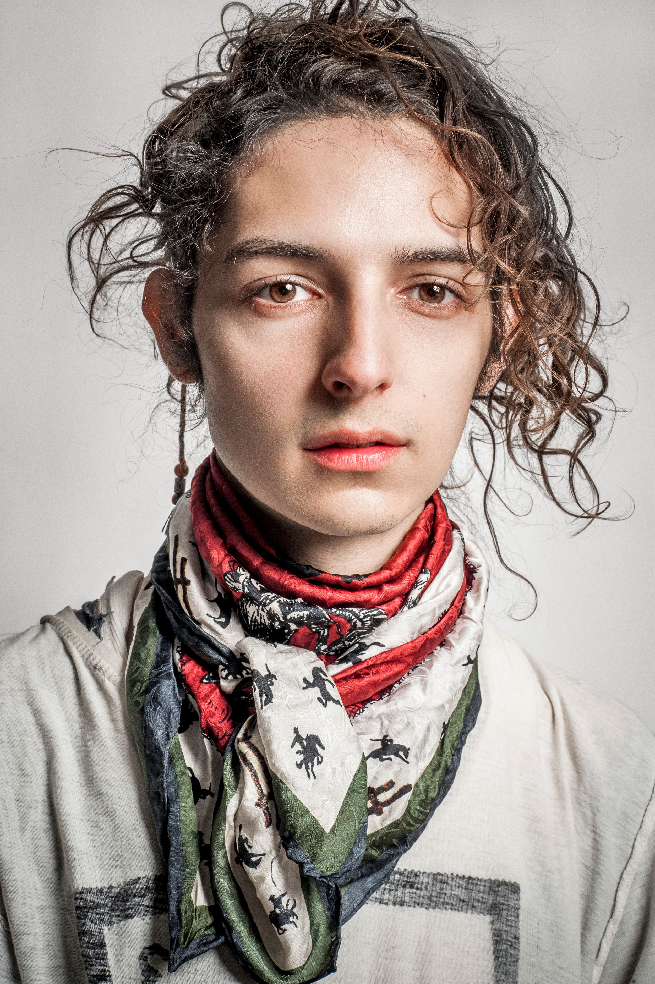 Young person with curly hair wearing a patterned scarf with red, white, and green colors against a gray background.
