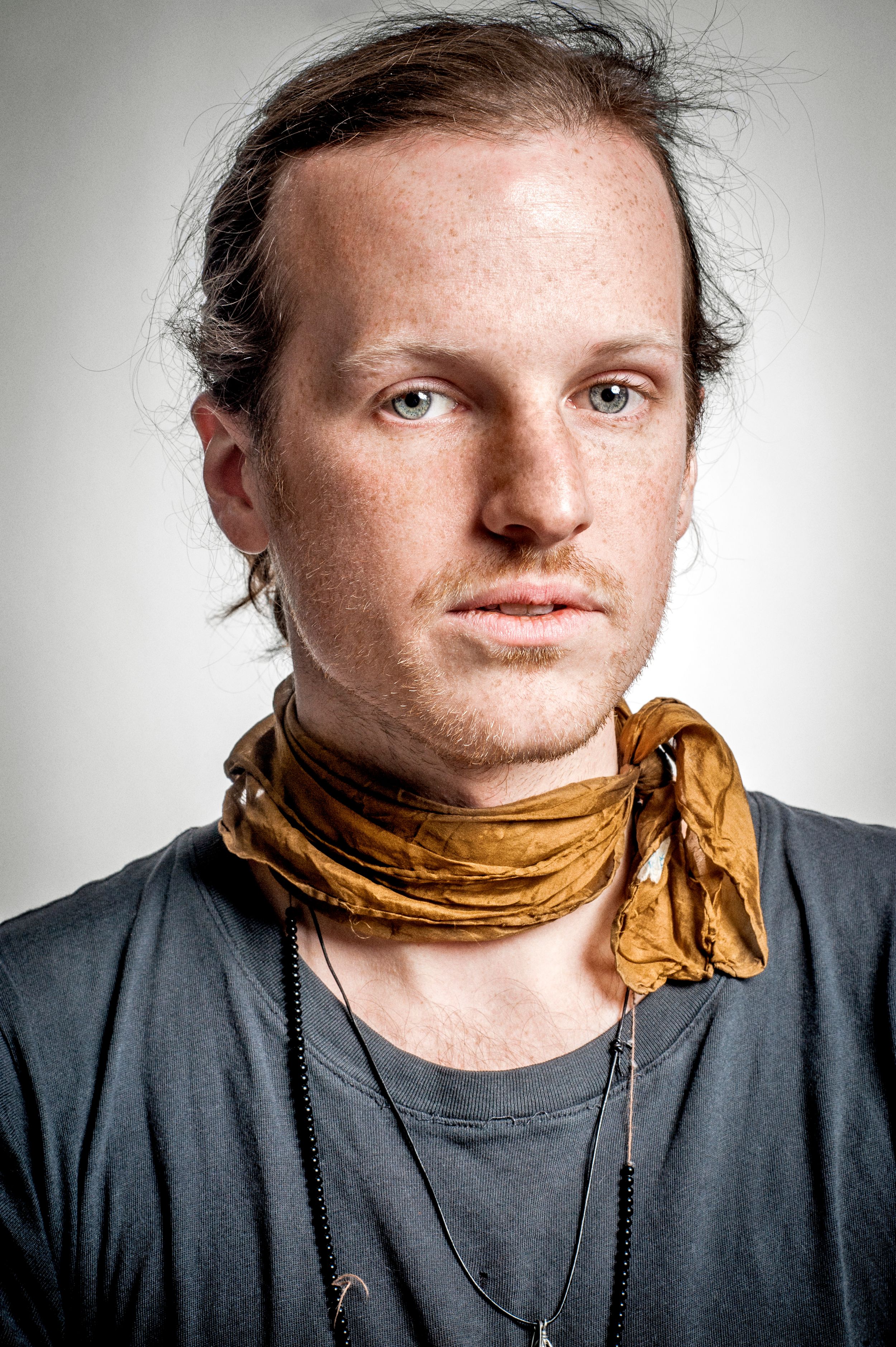 Young man with freckles wearing a gray t-shirt and golden yellow neck scarf with a chain necklace.