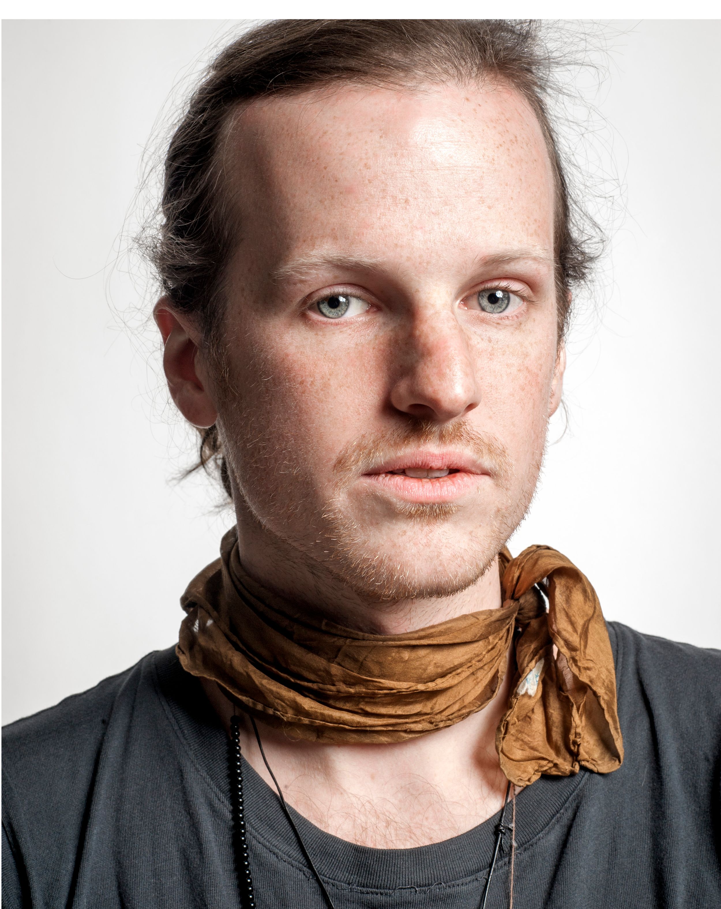 Young man with blue eyes, freckles, brown hair, wearing a tan neck scarf and dark shirt, portrait photo.
