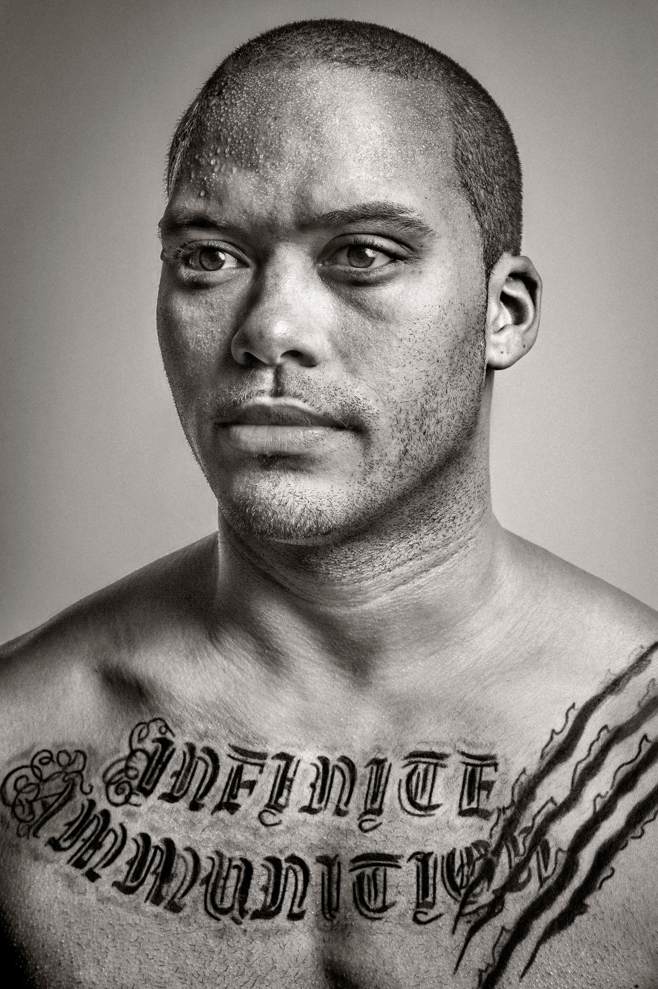 Black and white portrait of a shirtless man with chest tattoo reading Infinite Ammunition.