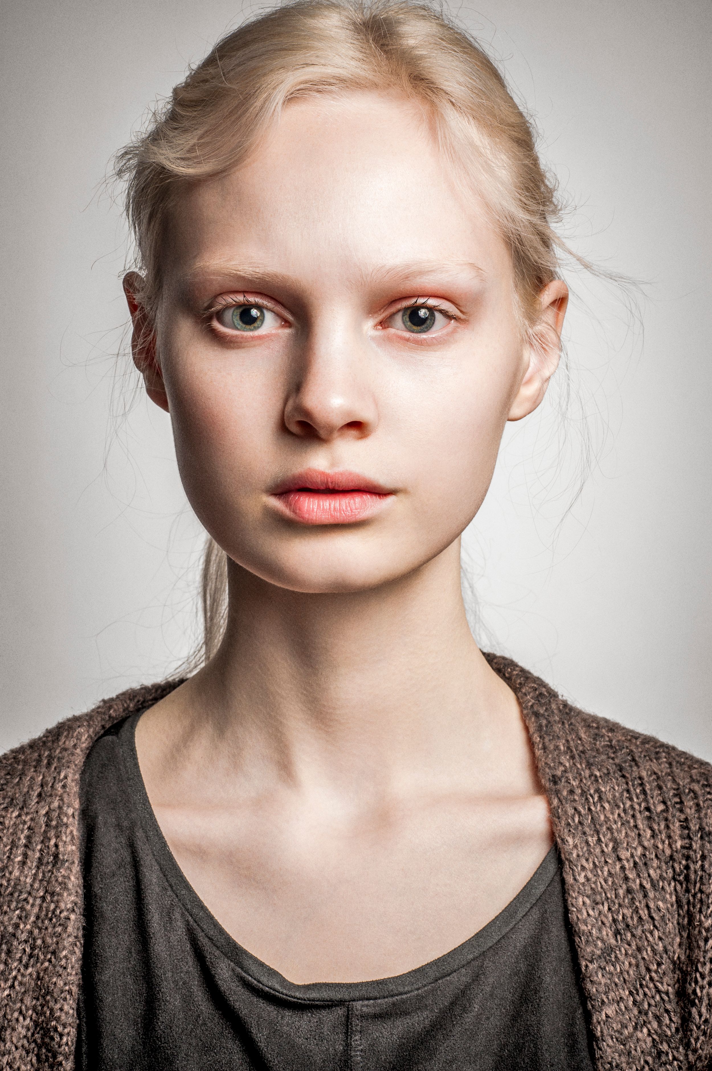 Young blonde woman with pale skin and minimal makeup wearing a gray top and brown knit cardigan against a light background.