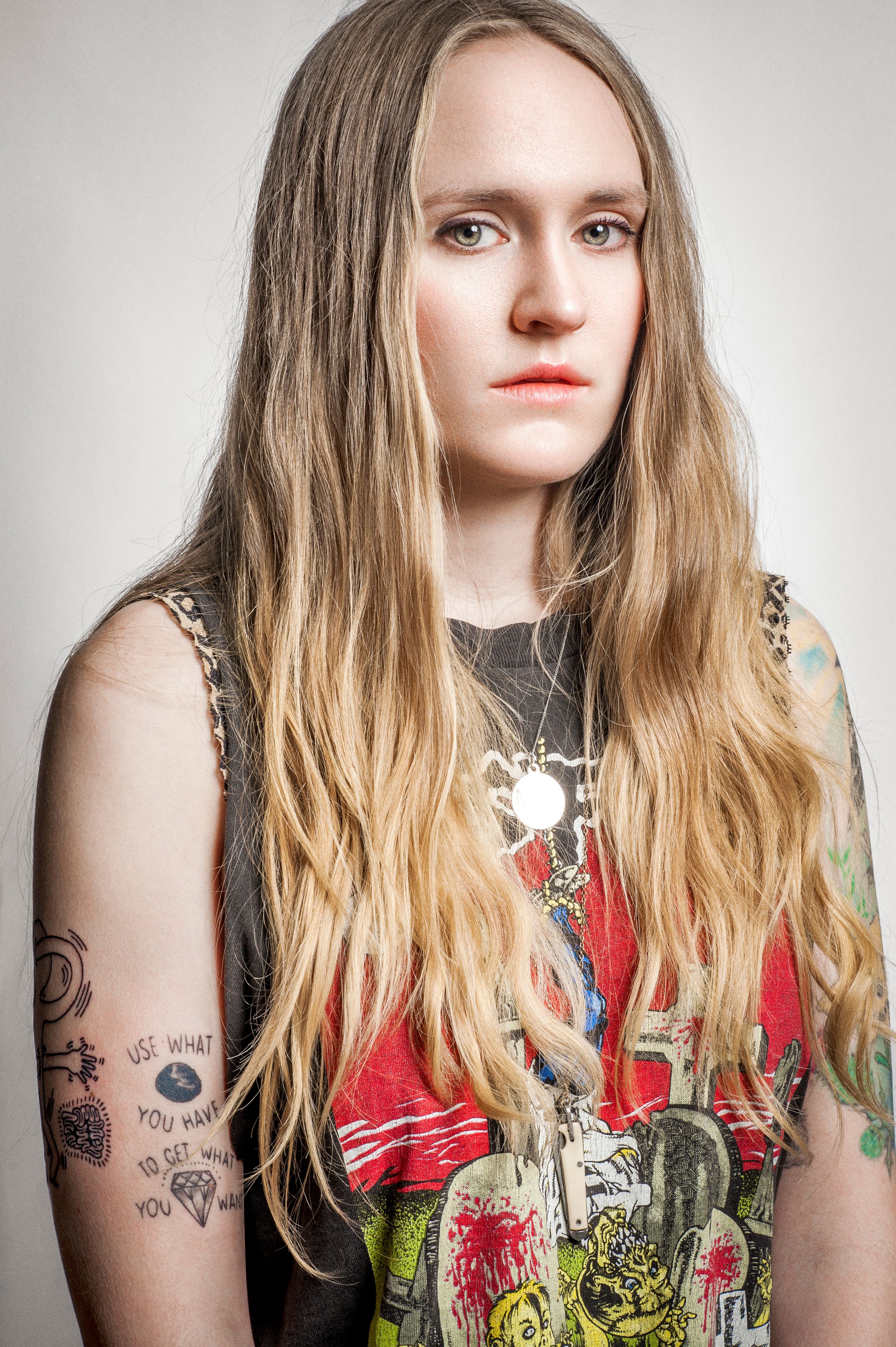 Young woman with long blonde hair, tattoos on arms, wearing colorful graphic tank top and layered necklaces.