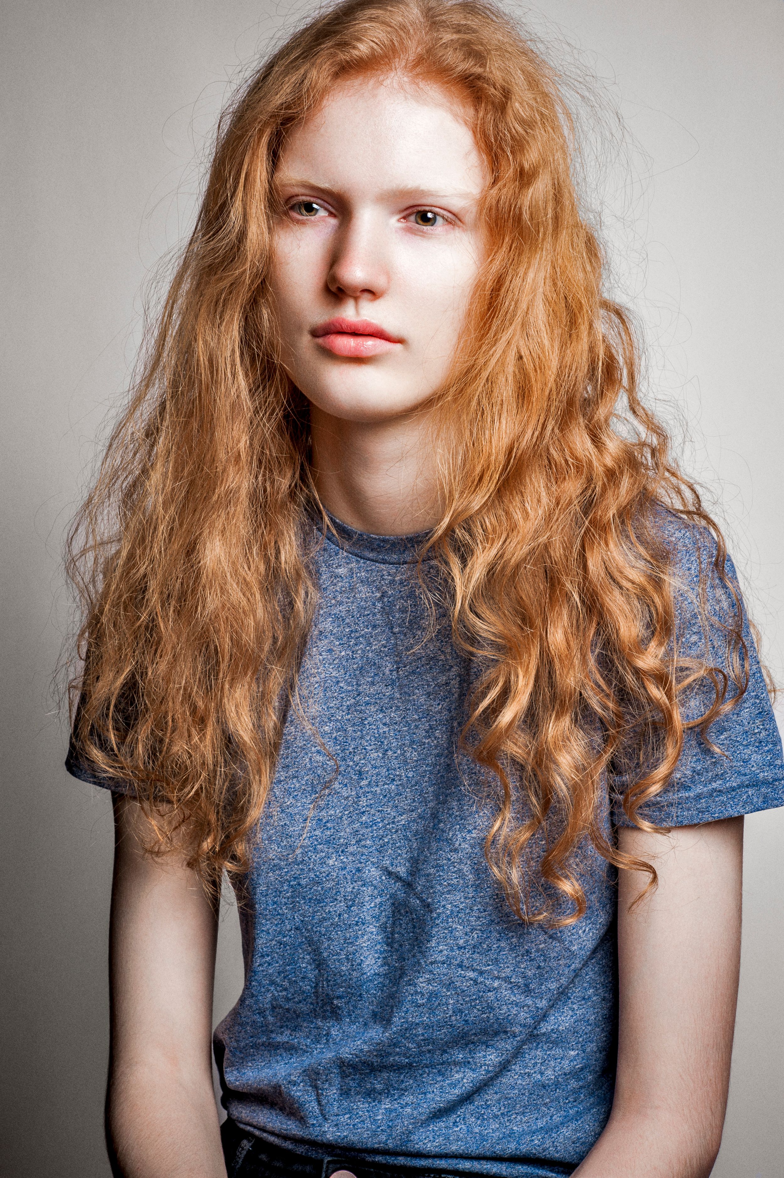Young woman with long curly red hair wearing a gray t-shirt poses against a light gray background.