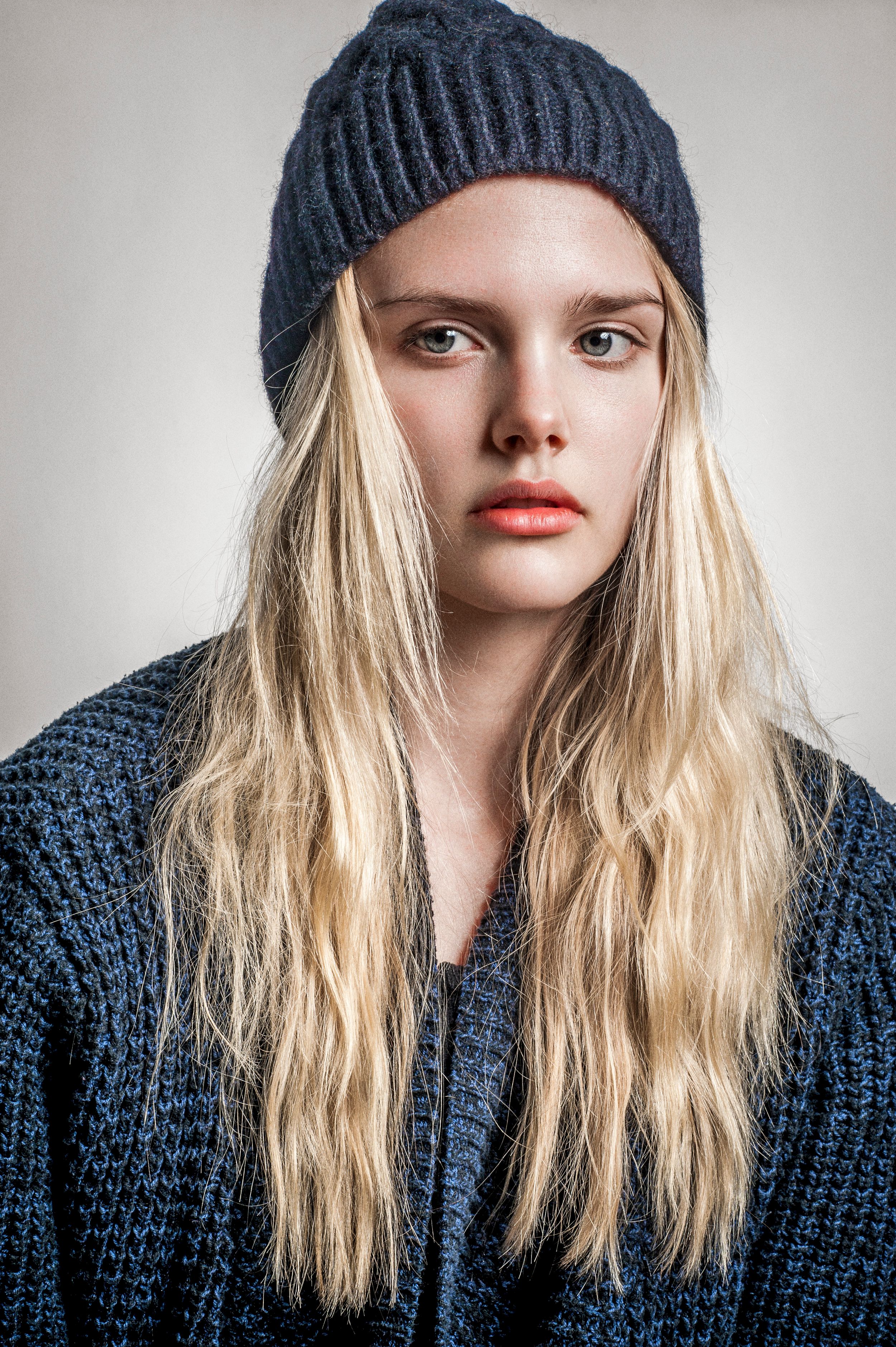 Young blonde woman wearing a dark knit beanie hat and blue cardigan sweater, studio portrait photography.
