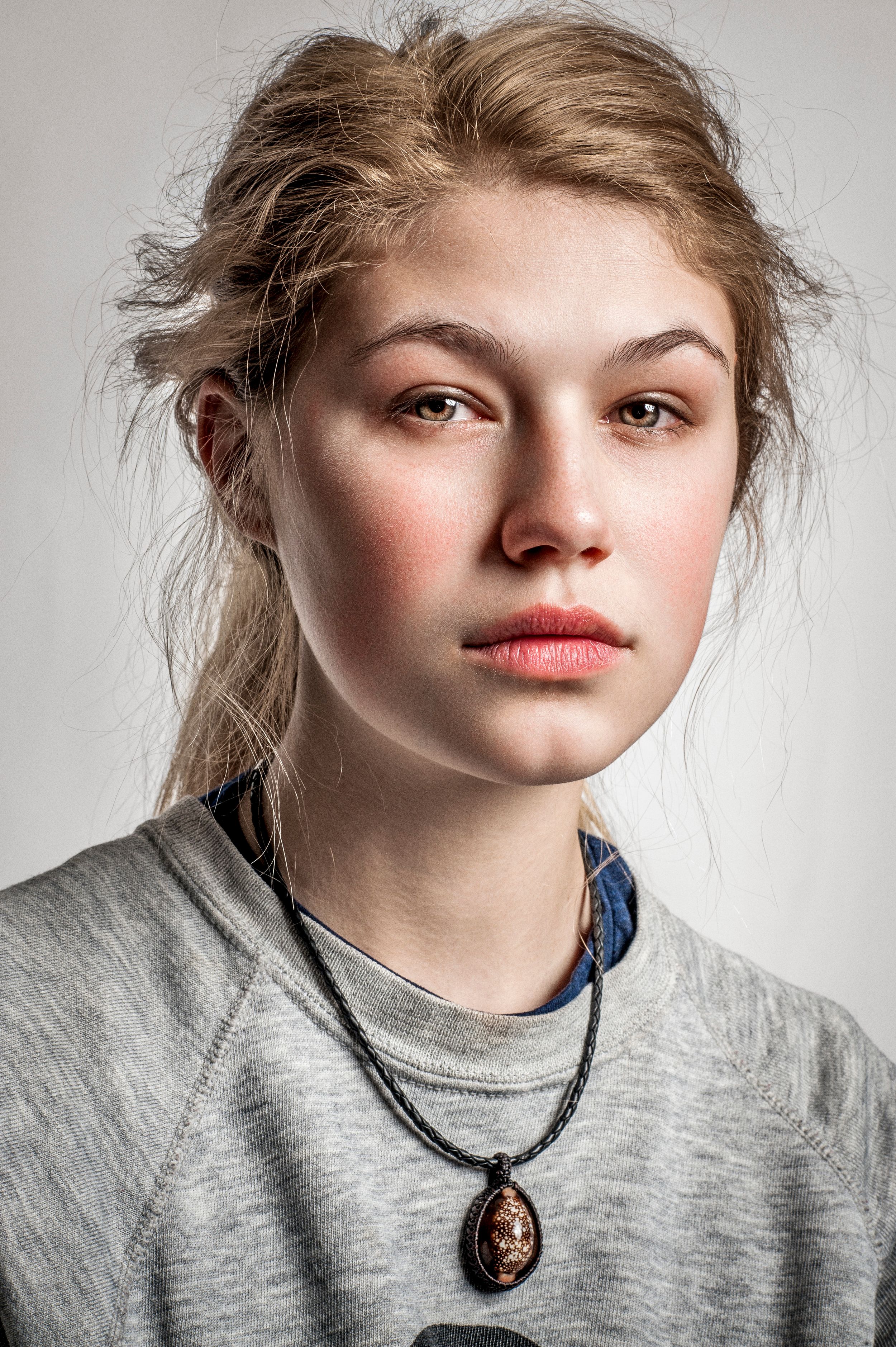 Young woman with blonde hair, rosy cheeks, and a pendant necklace wearing a gray sweatshirt against a light background.