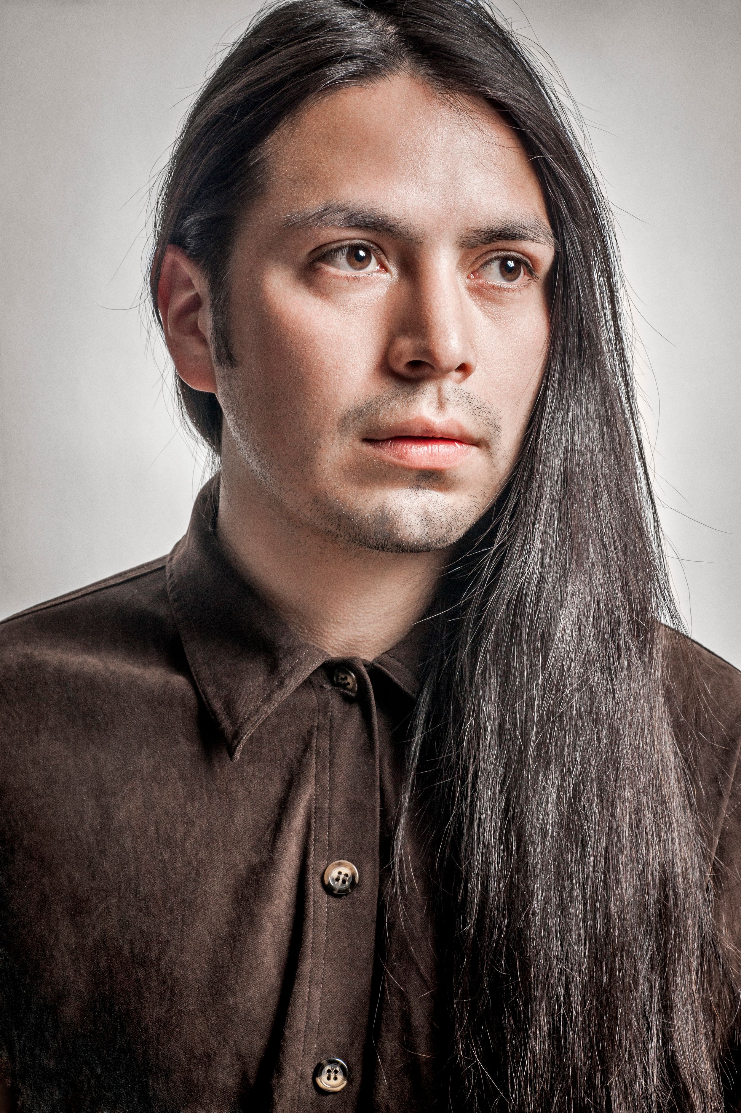 Young man with long dark hair wearing a brown button-up shirt, portrait studio photo.