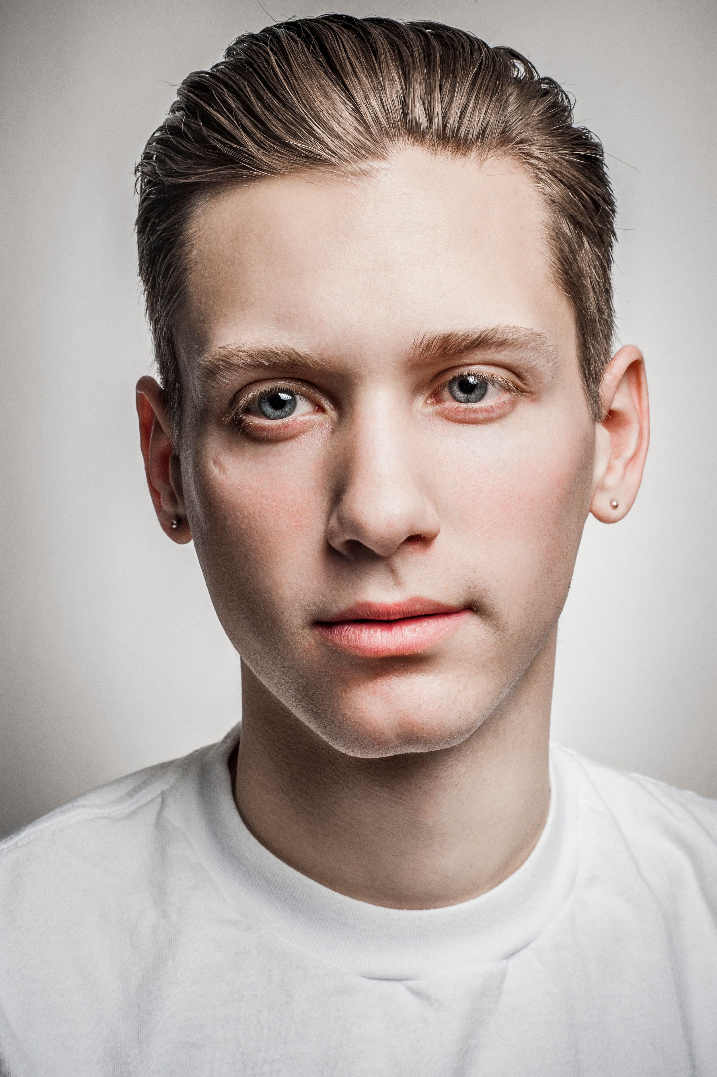 Young man with slicked-back hair and small ear studs wearing a white t-shirt against a light gray background.