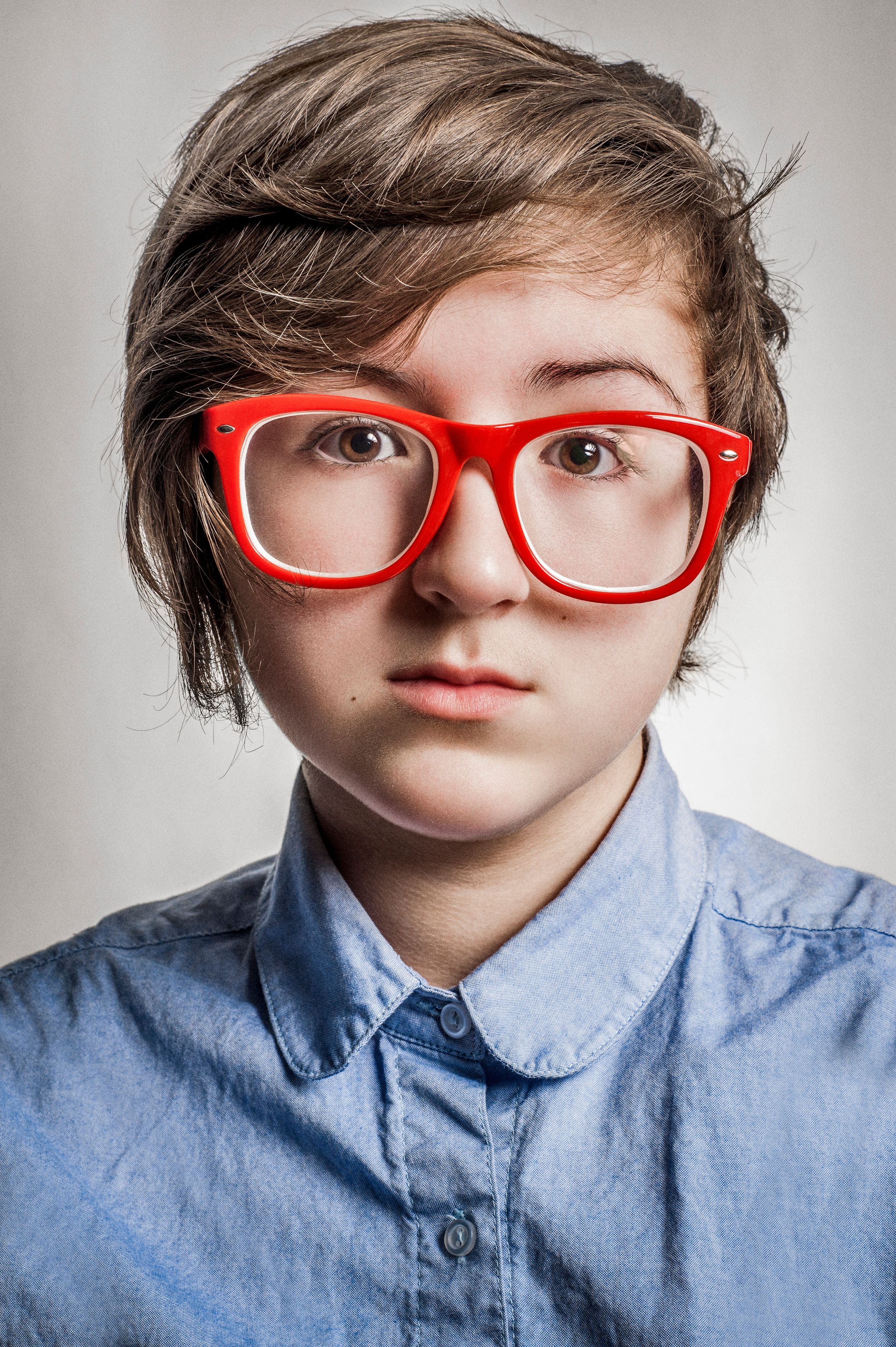 Young person with short brown hair wearing oversized red glasses and a blue chambray shirt against gray background.