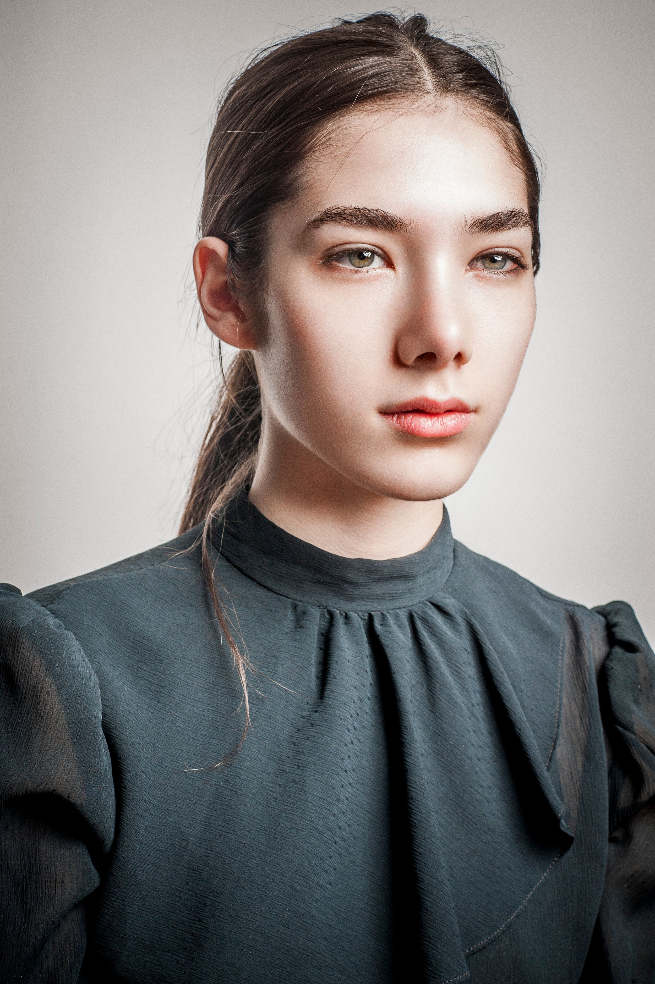 Young woman with dark hair in ponytail wearing teal high-neck blouse with puff sleeves, studio portrait.