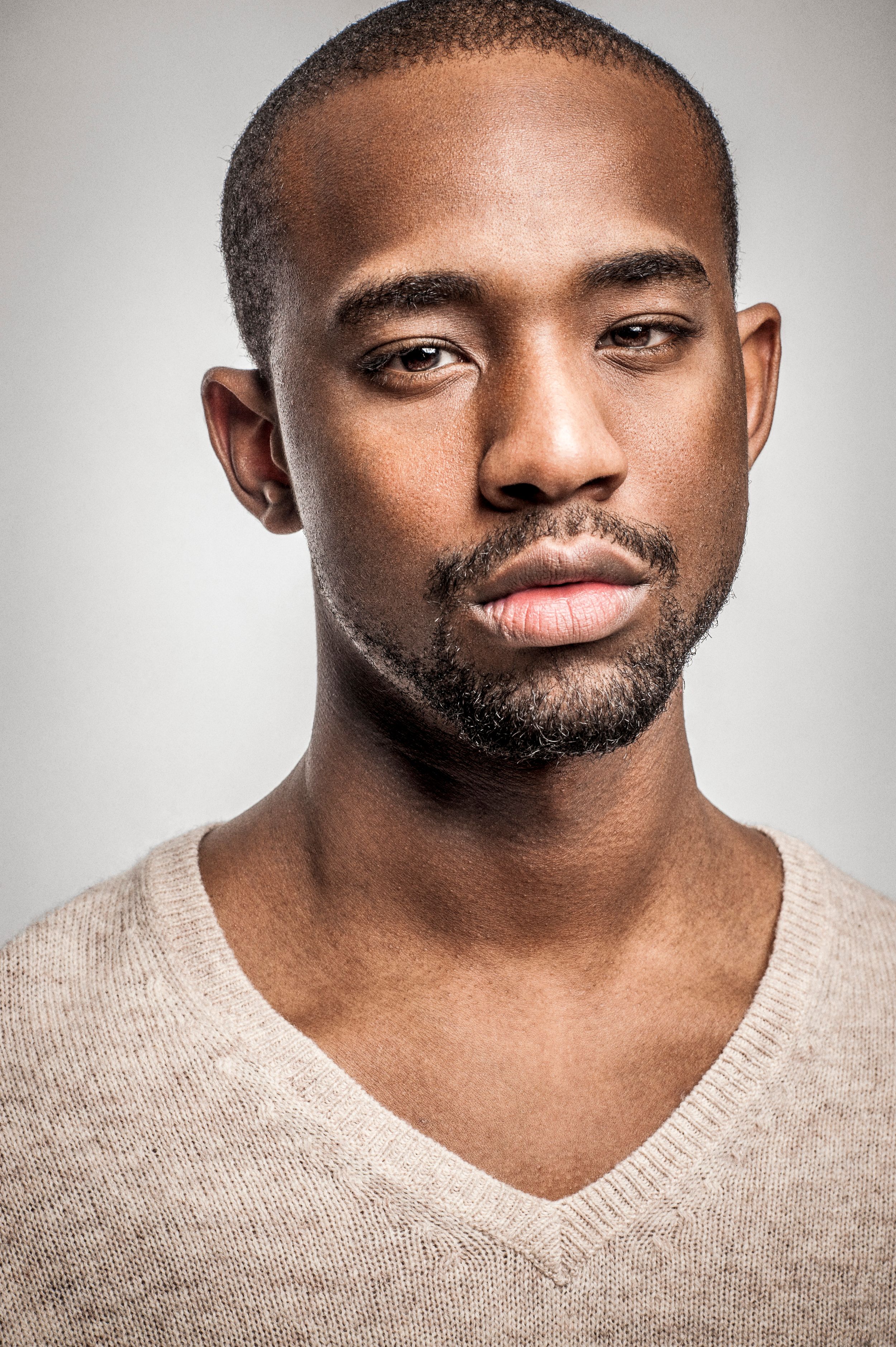Close-up portrait of a young Black man with short hair and beard wearing a beige v-neck sweater on gray background.