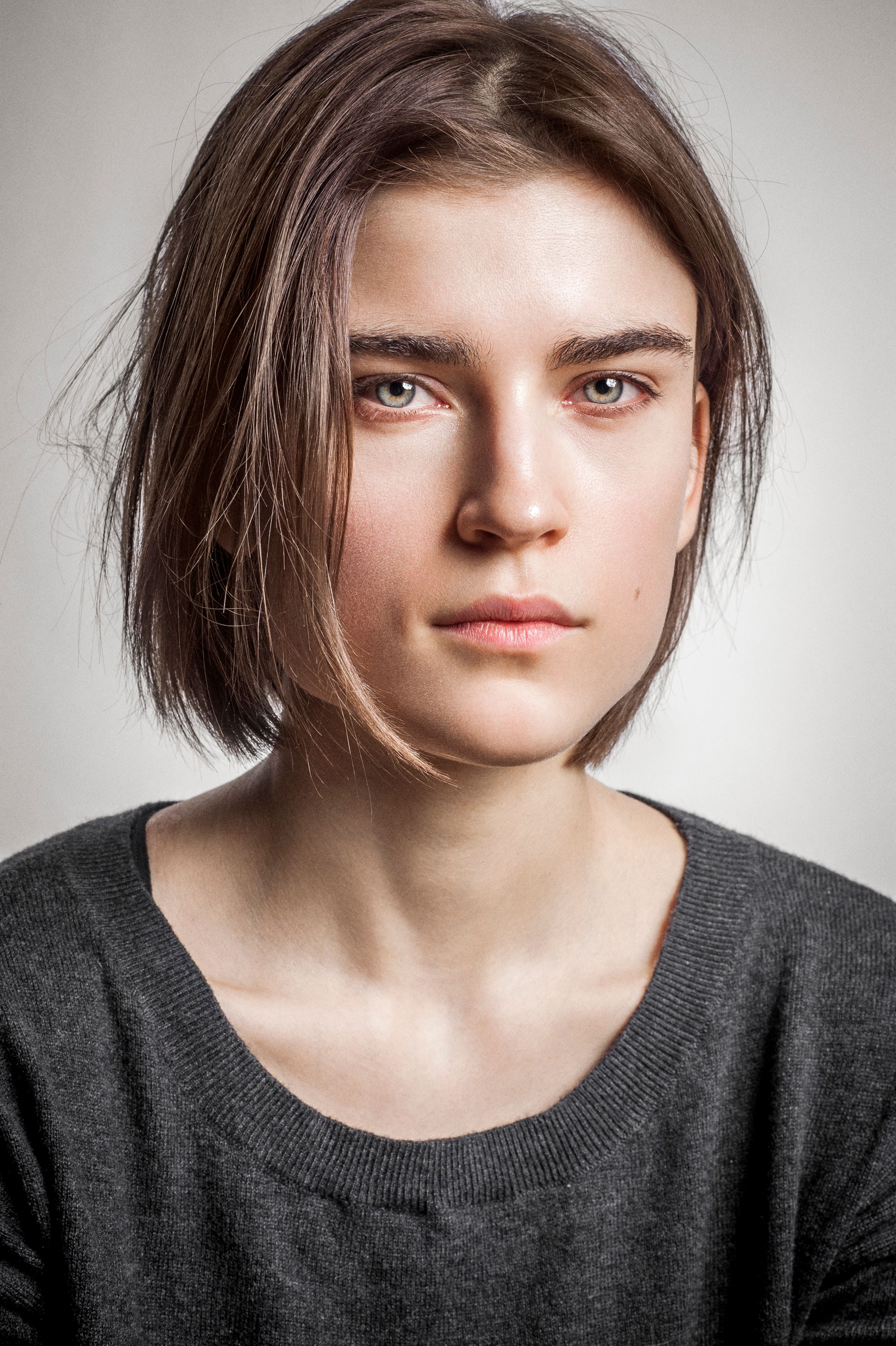 Young woman with short brown hair and serious expression wearing a gray sweater against a light background.