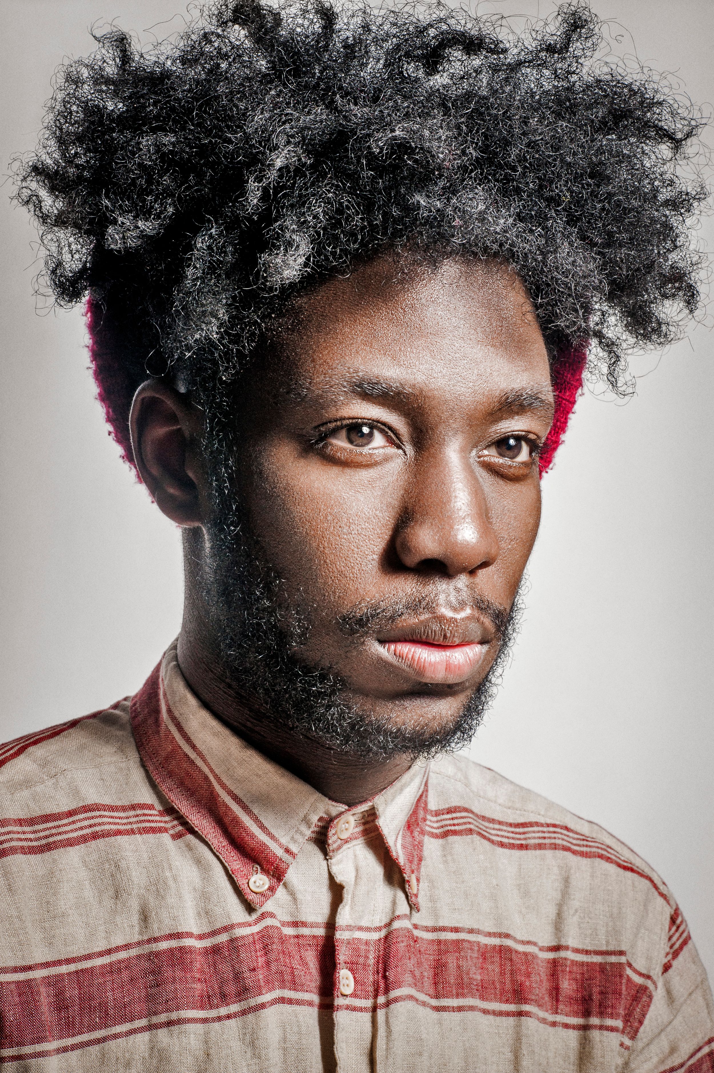 Young Black man with natural afro hair and beard wearing a striped linen shirt, portrait photo on gray background.