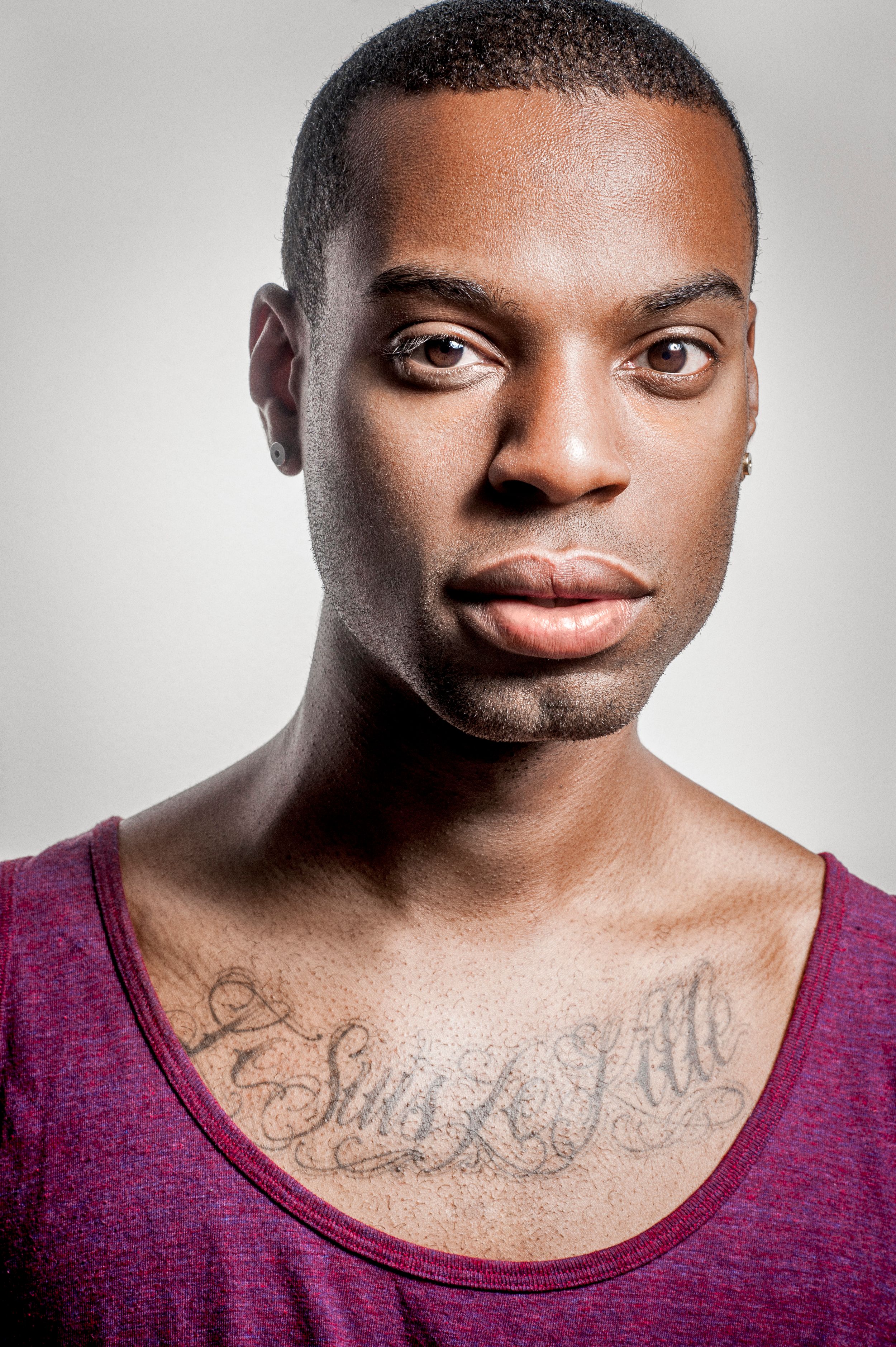 Young Black man with chest tattoo reading Jesus Kills wearing a red tank top against a gray background.