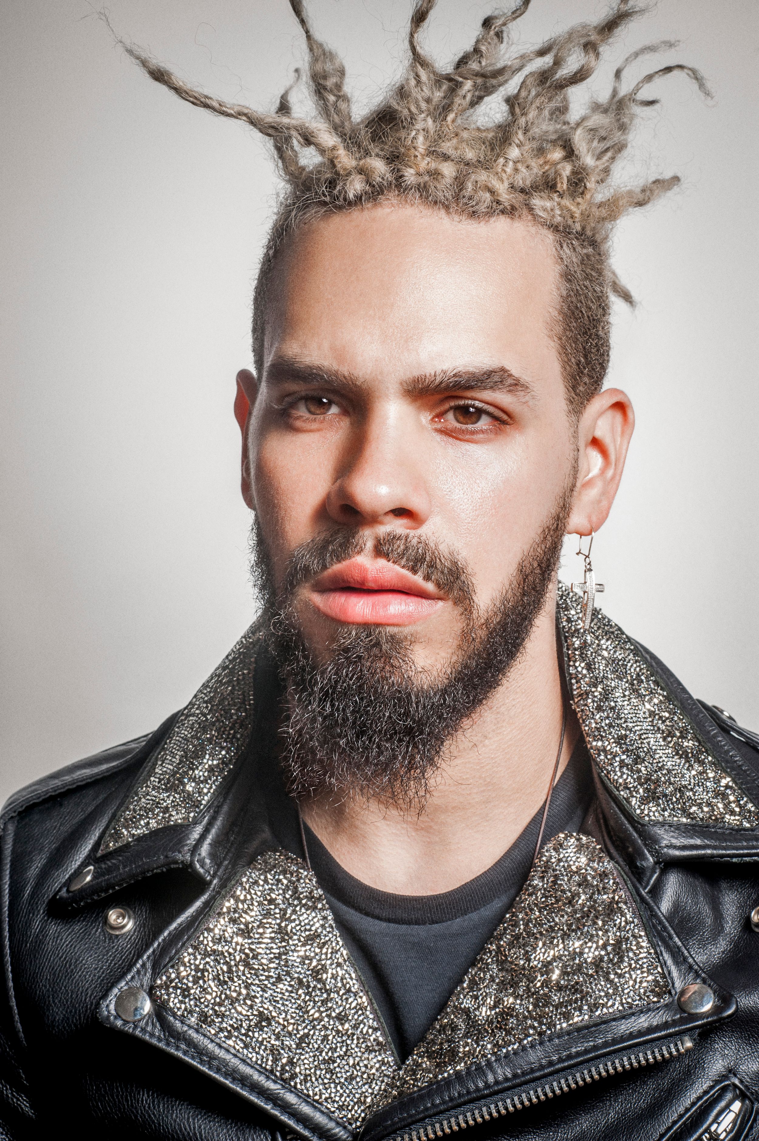 Man with blonde dreadlocks, beard, earring, wearing a glittery black leather jacket, posing against a light background.