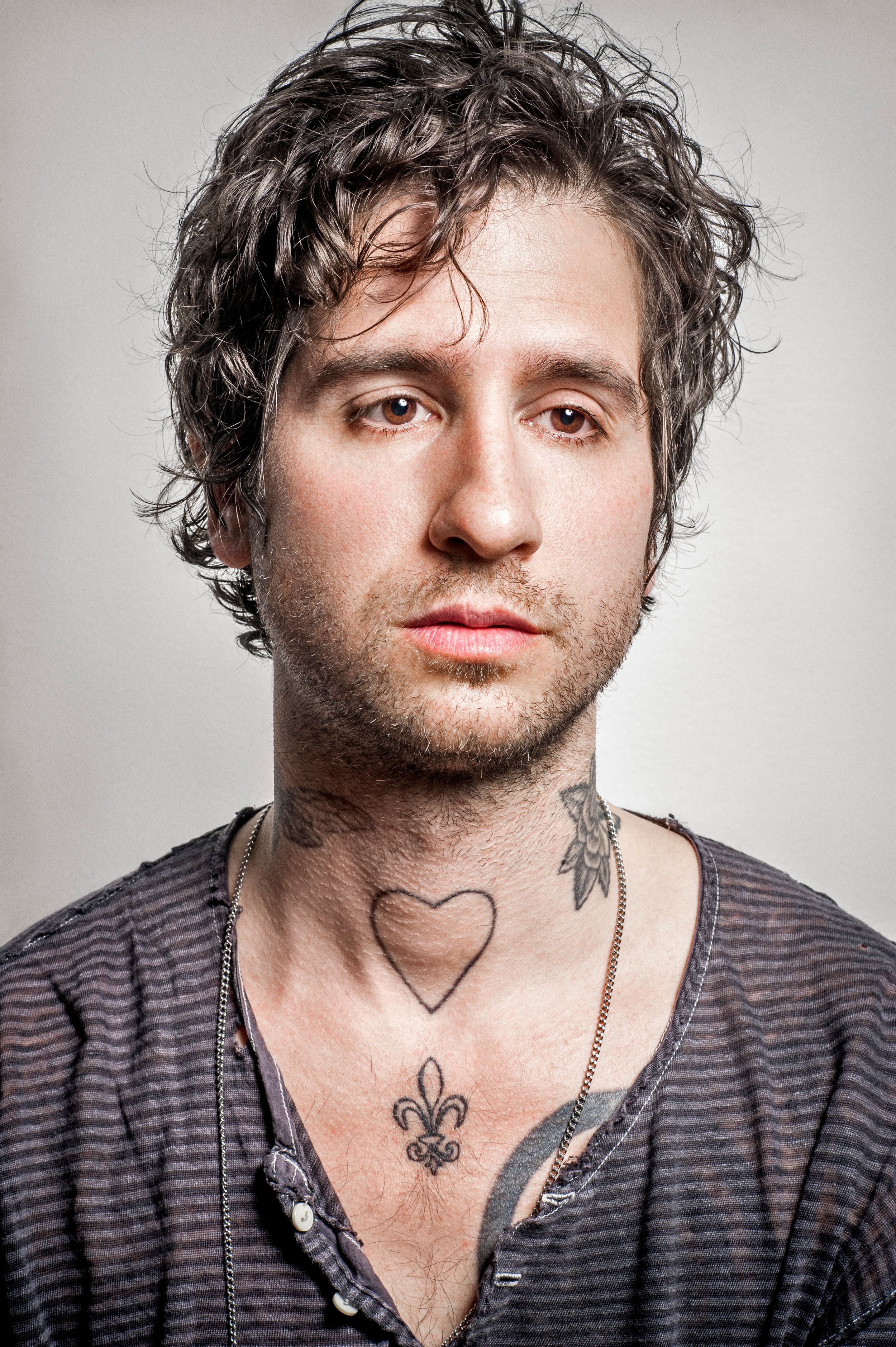 Young man with curly hair, heart and fleur-de-lis chest tattoos, wearing a striped V-neck shirt, posing against gray background.