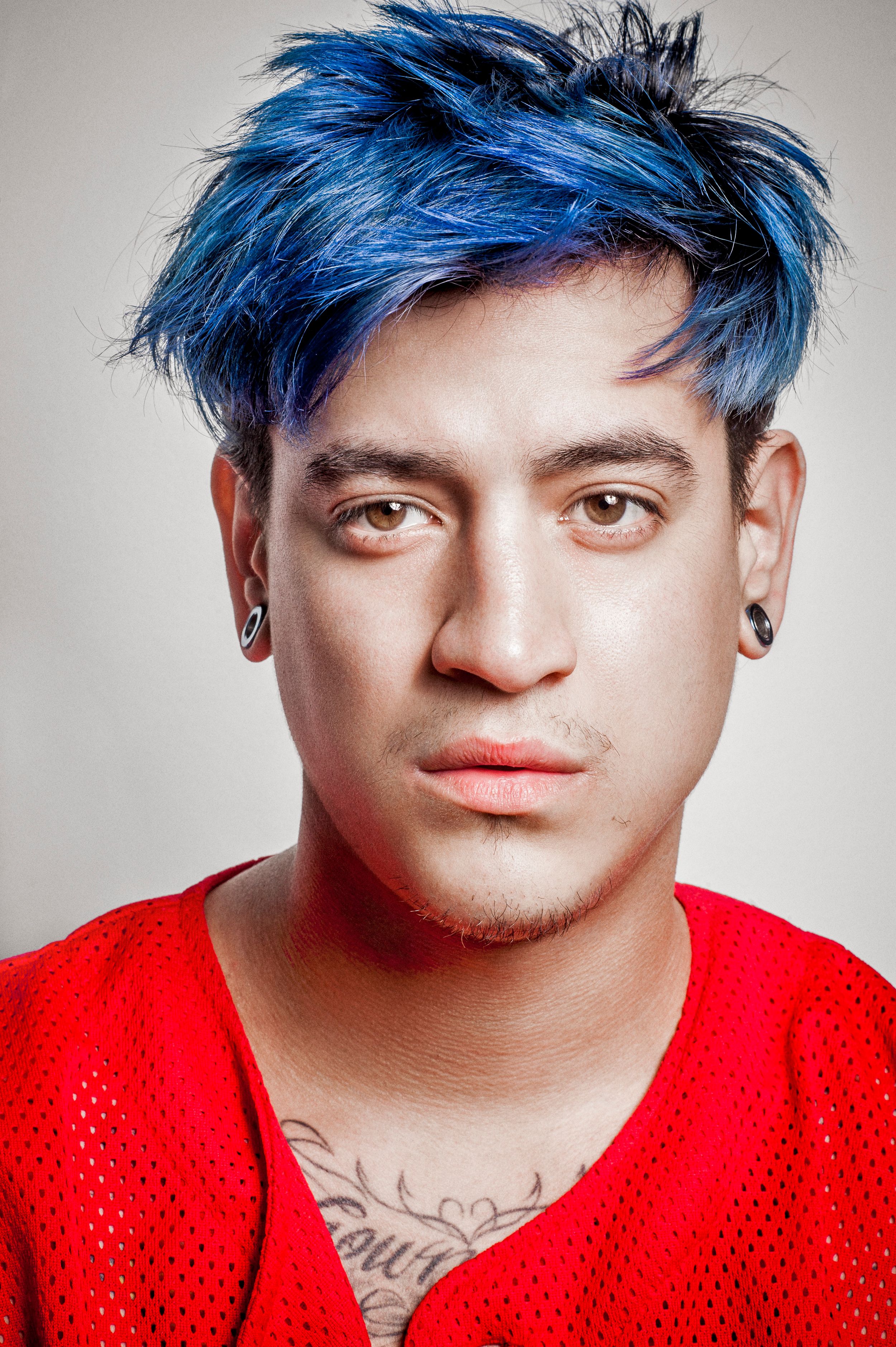 Young man with vibrant blue hair, ear gauges, neck tattoo, wearing a red mesh shirt against a gray background.