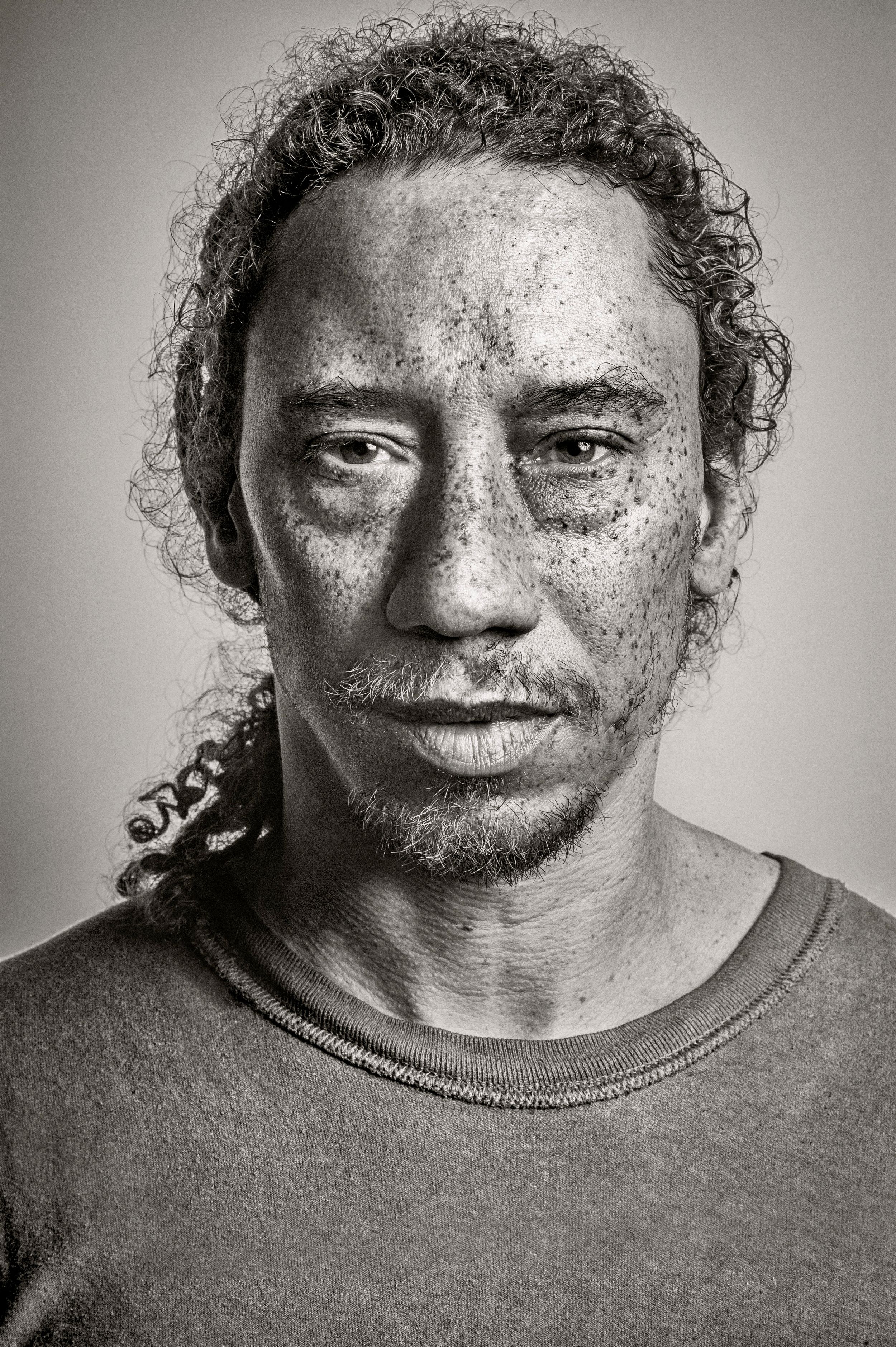 Black and white portrait of a man with curly hair, freckles, and a chain necklace wearing a crew neck shirt.
