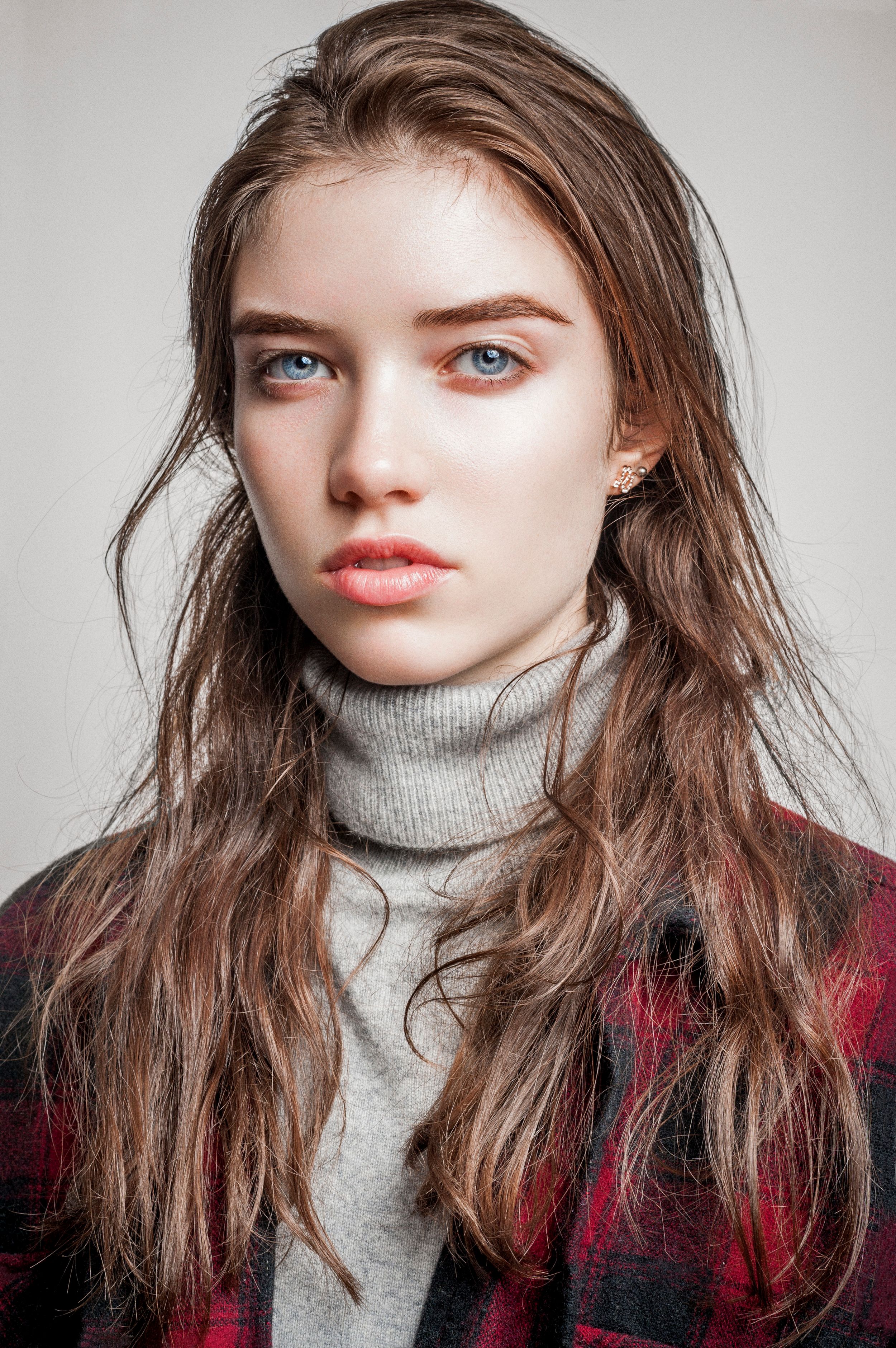 Young woman with long brown hair and blue eyes wearing a gray turtleneck and plaid jacket poses for portrait.