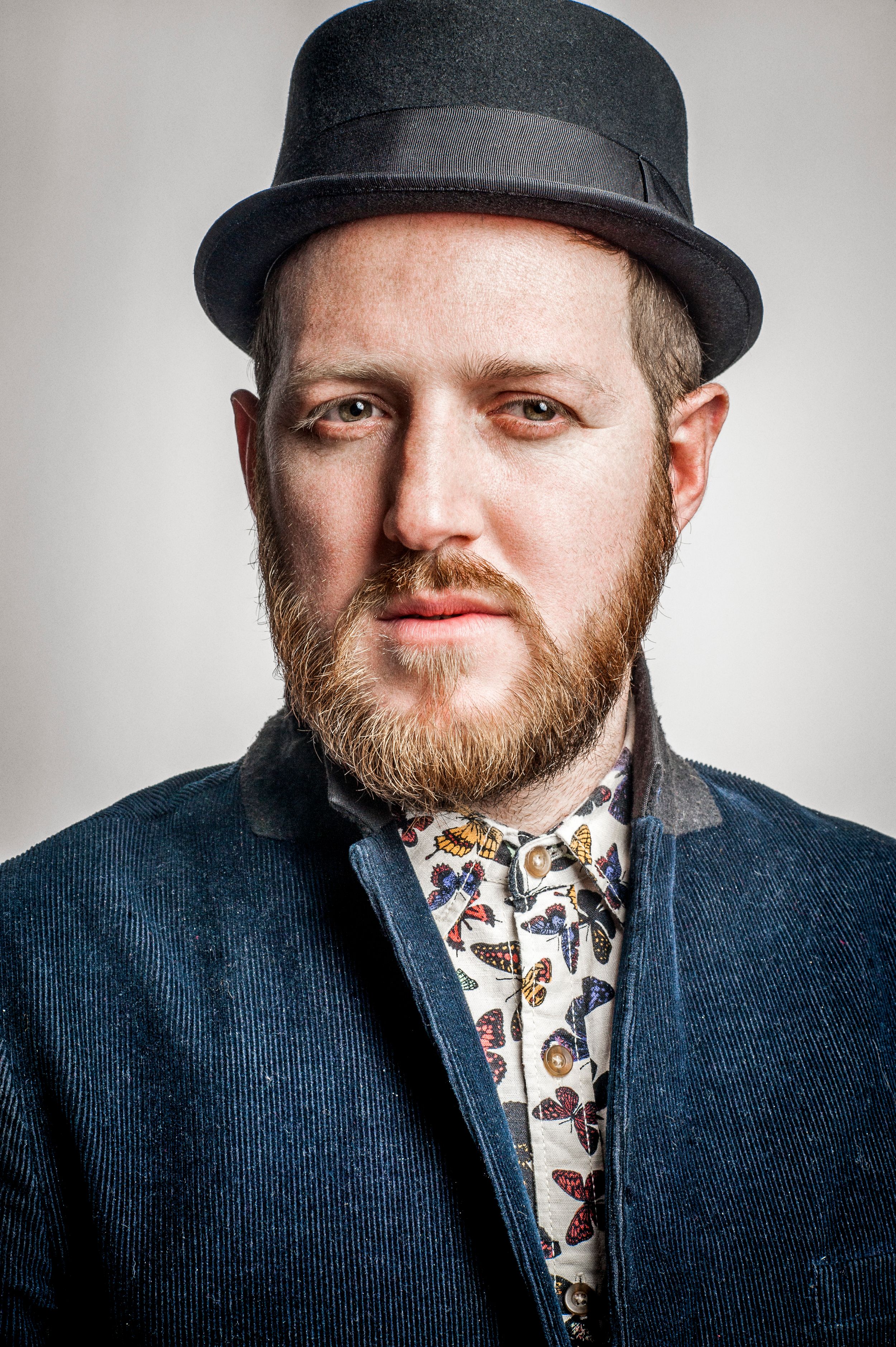 Bearded man wearing a black fedora hat and patterned shirt under a blue blazer, posing for a portrait photo.