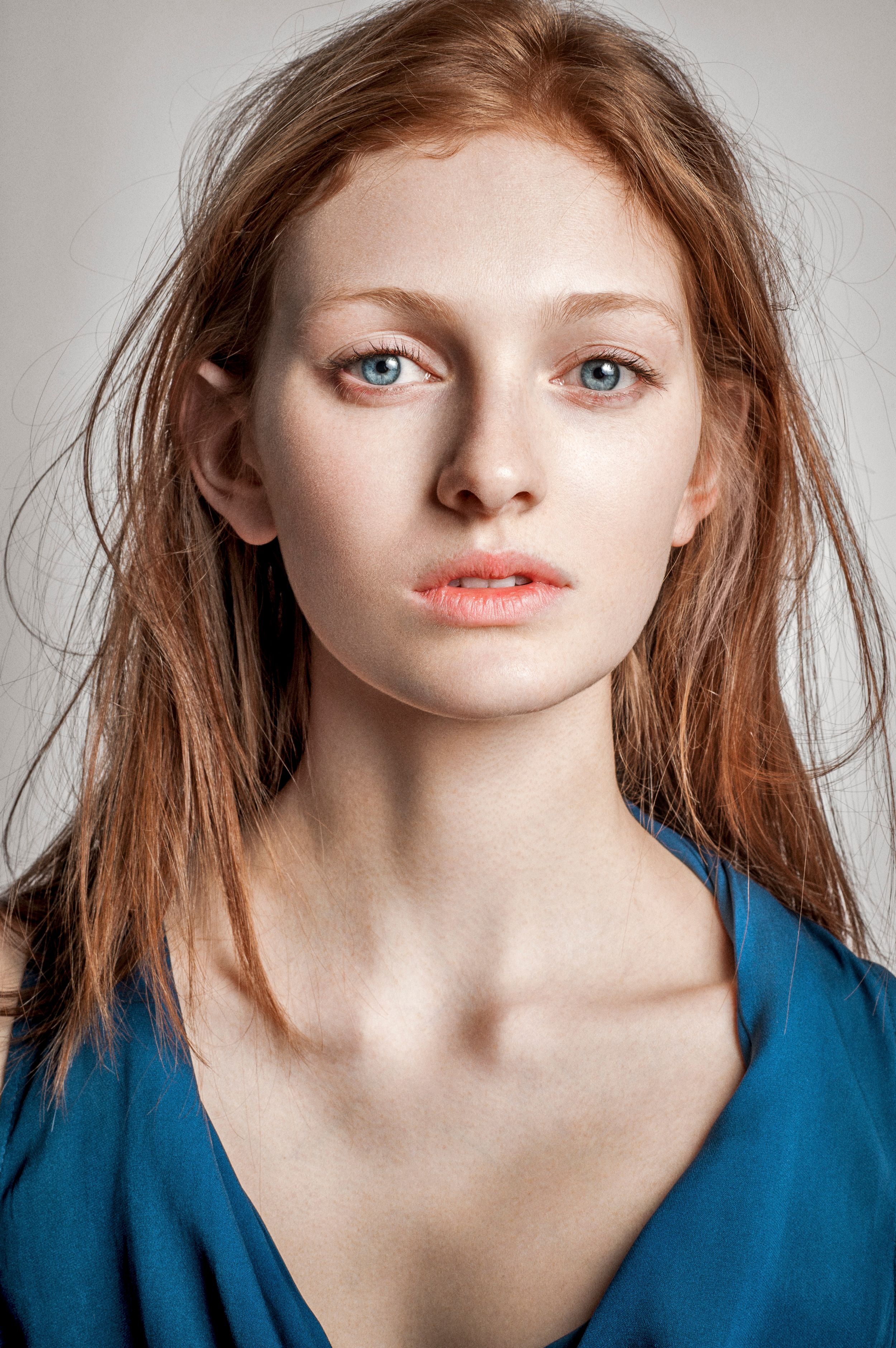 Young woman with red hair and blue eyes wearing a teal top, posing for a portrait against a light background.