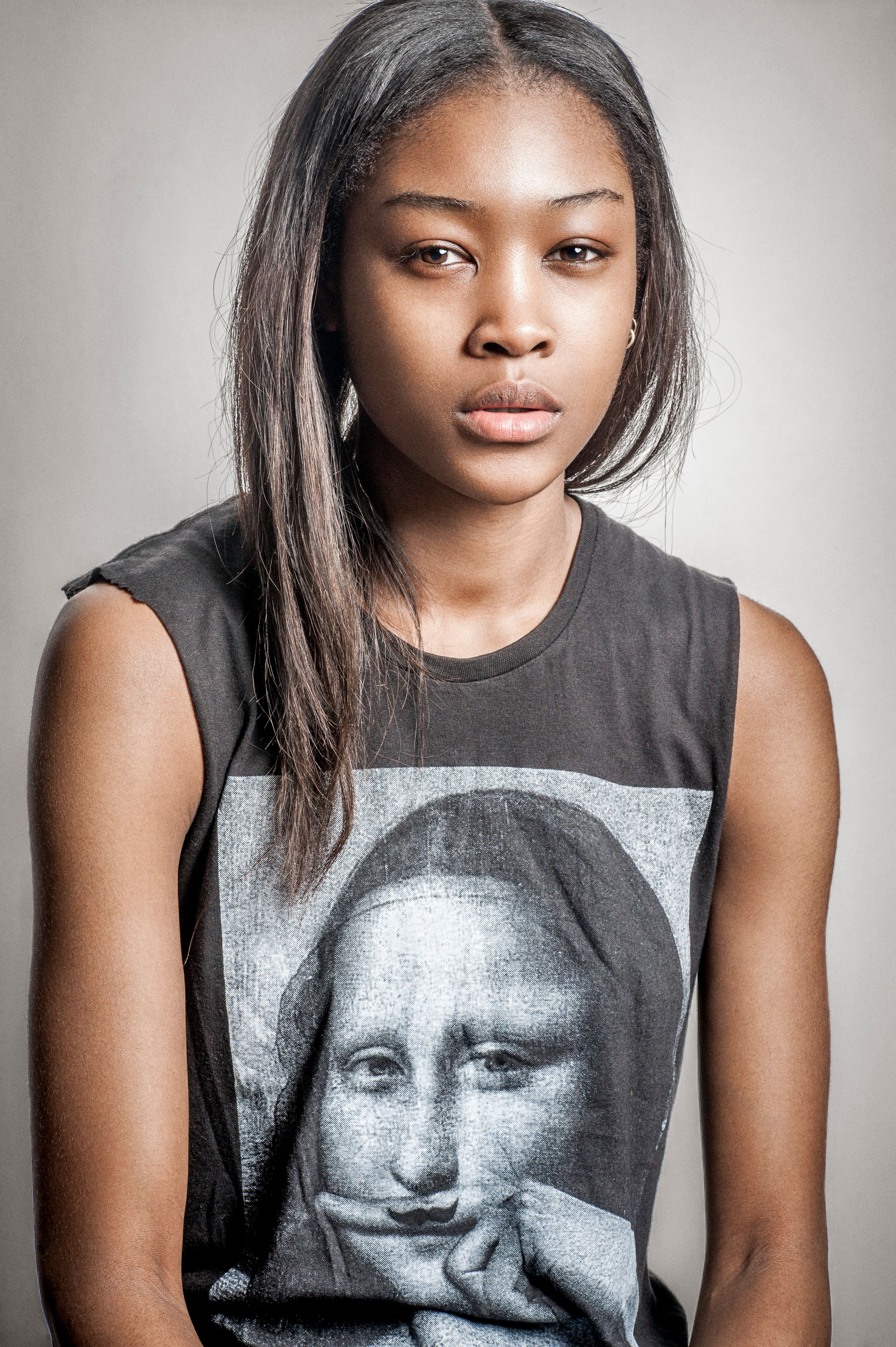 Young woman with long straight hair wearing a black sleeveless Mona Lisa graphic tee, posing against a gray background.