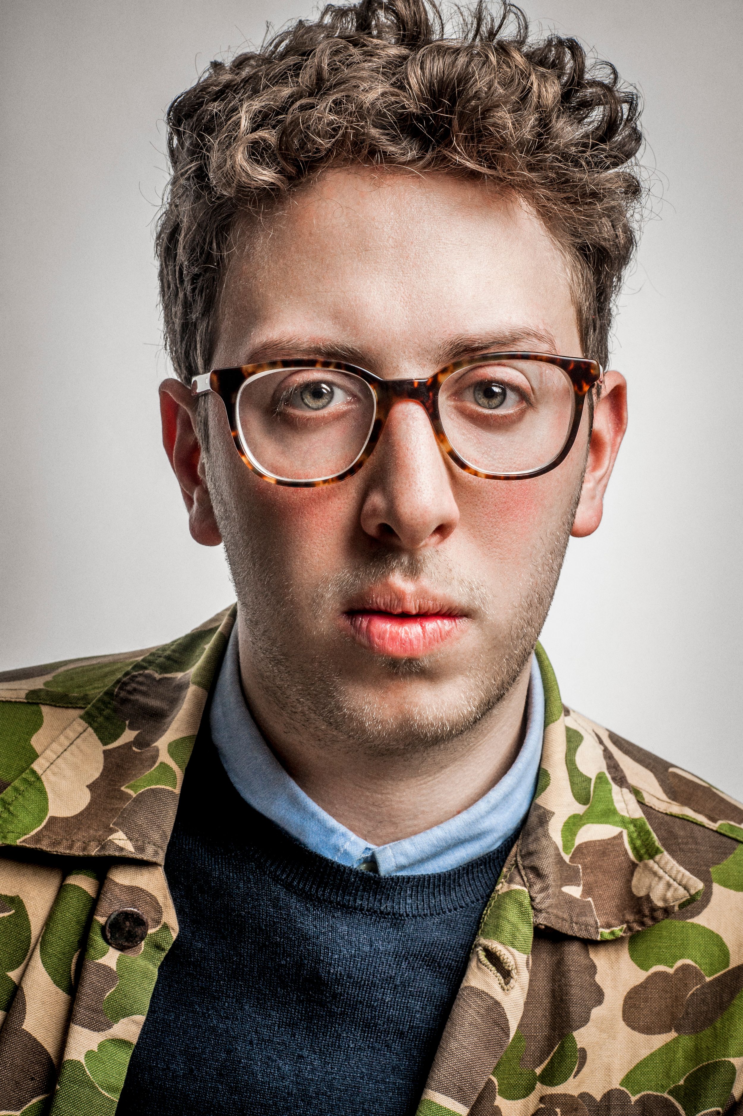 Young man with curly hair and tortoiseshell glasses wearing a camouflage jacket over a dark sweater.