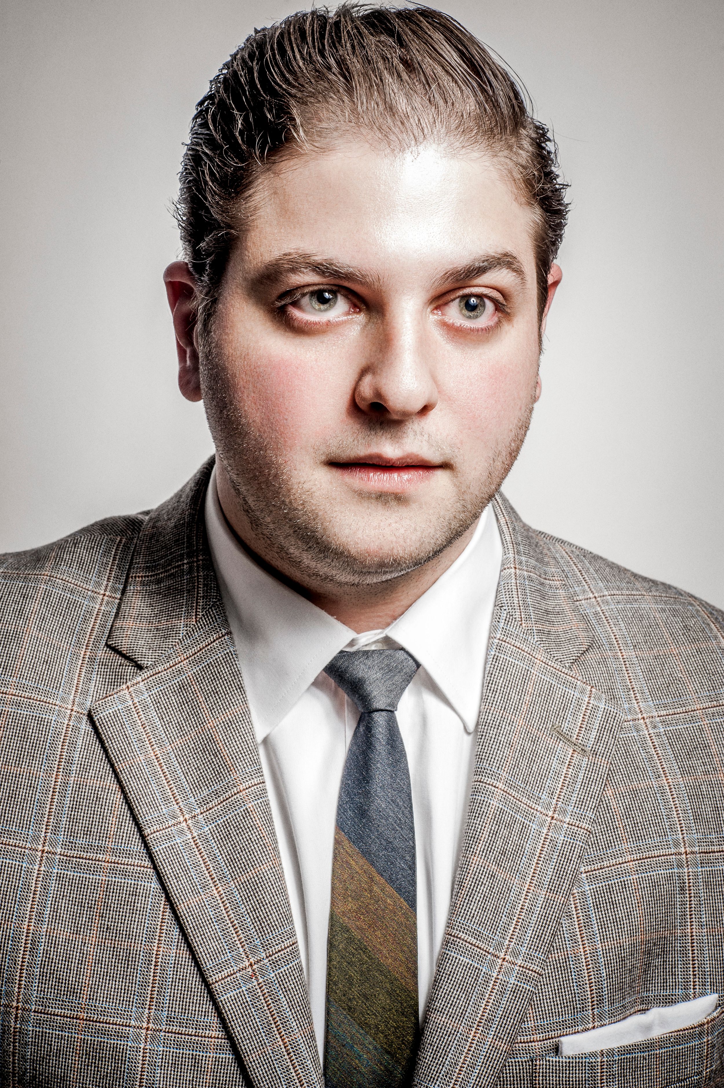 Man in gray plaid suit with tie and white pocket square posing for a professional portrait.