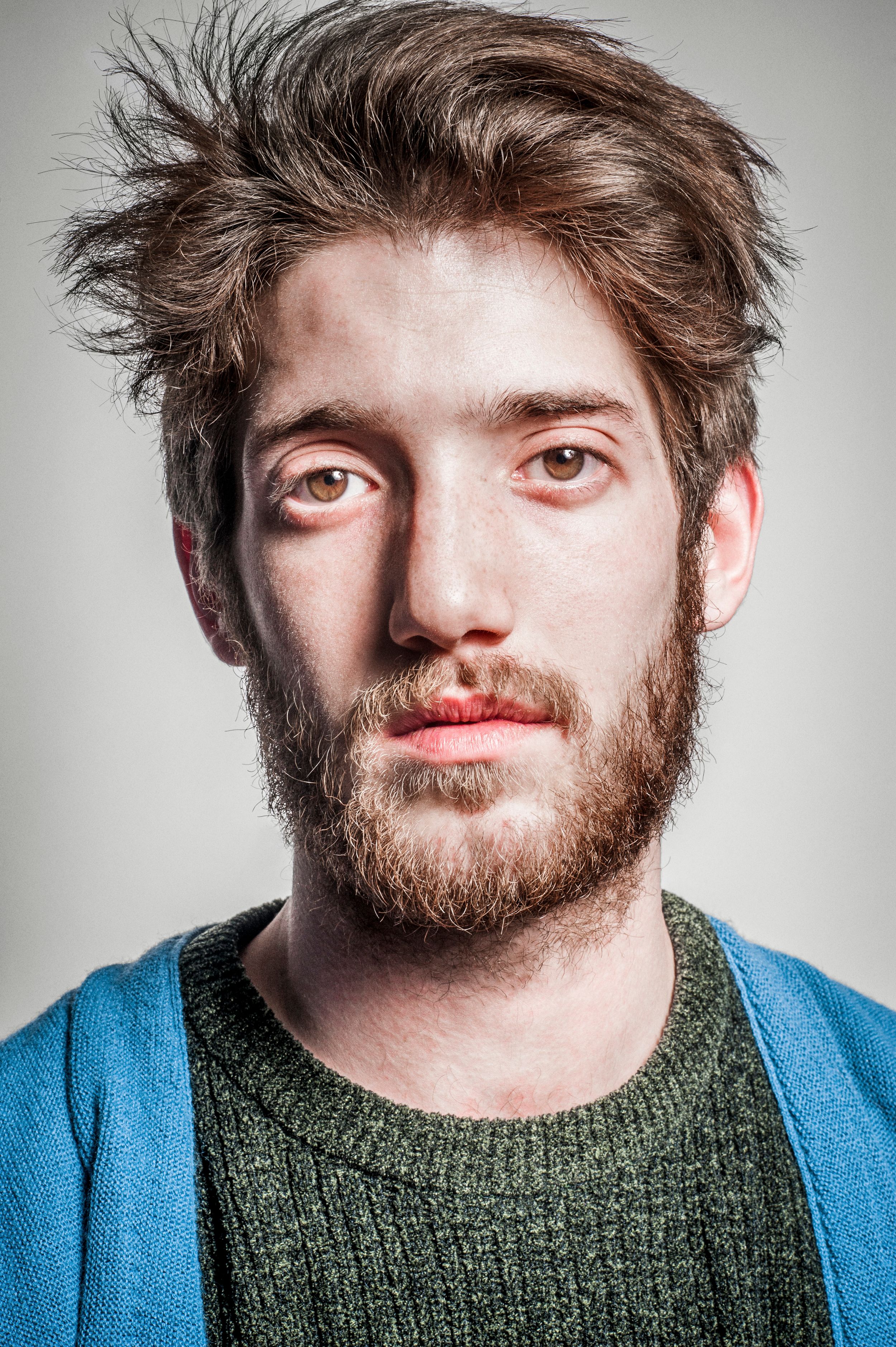 Young bearded man with messy brown hair wearing a green sweater and blue cardigan against a gray background.