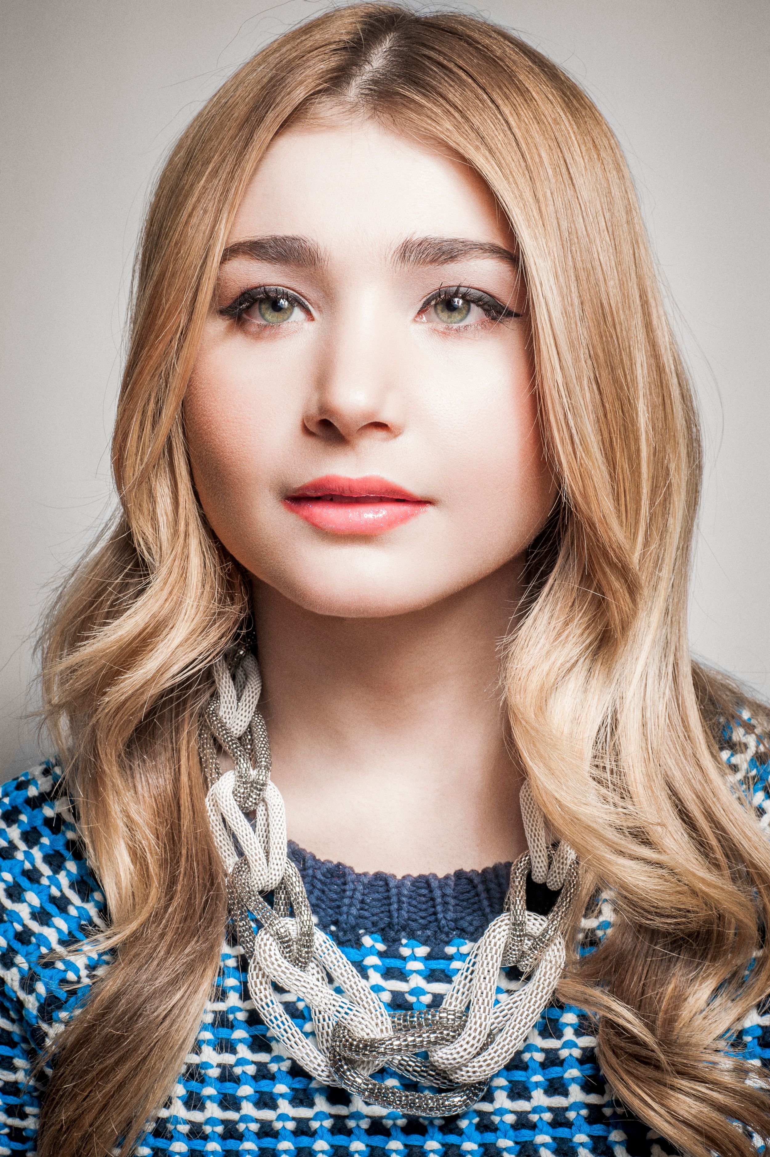 Young woman with long blonde hair wearing a blue knit sweater and silver chain necklace, posing for a portrait.