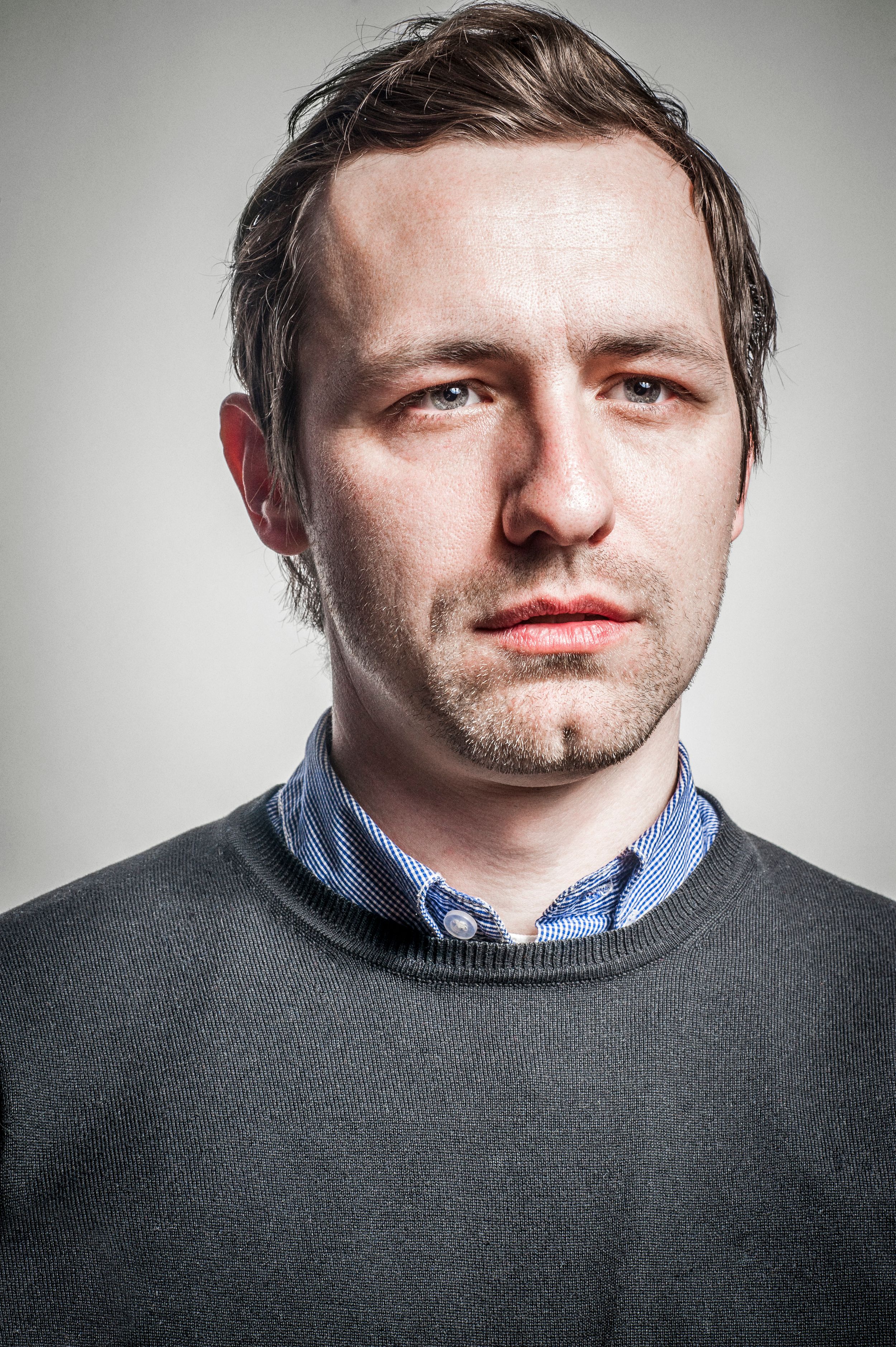 Portrait of a serious young man with stubble wearing a gray sweater over a blue collared shirt against a gray background.