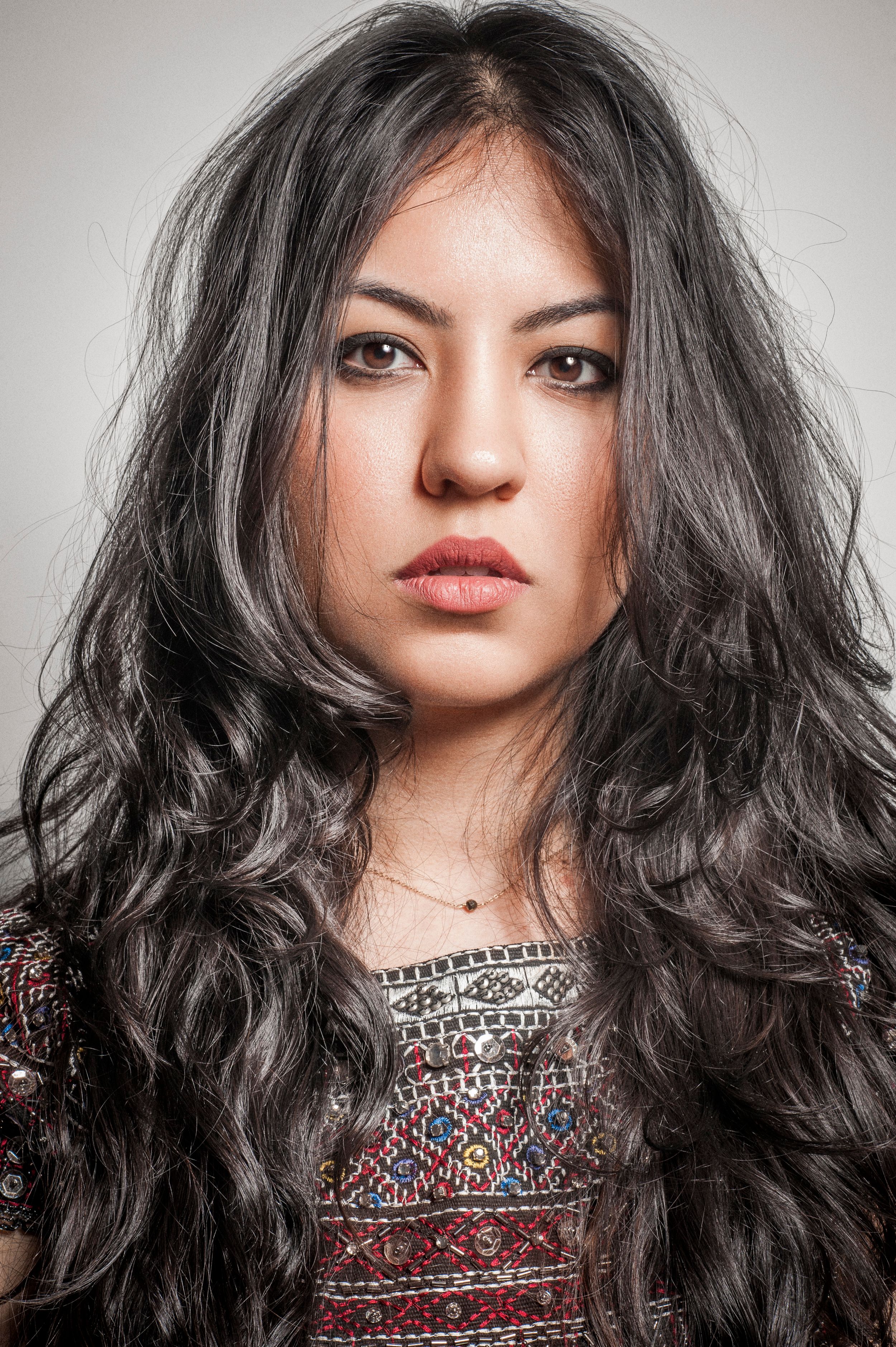 Young woman with long, dark wavy hair wearing a colorful patterned top, posing against a light gray background.