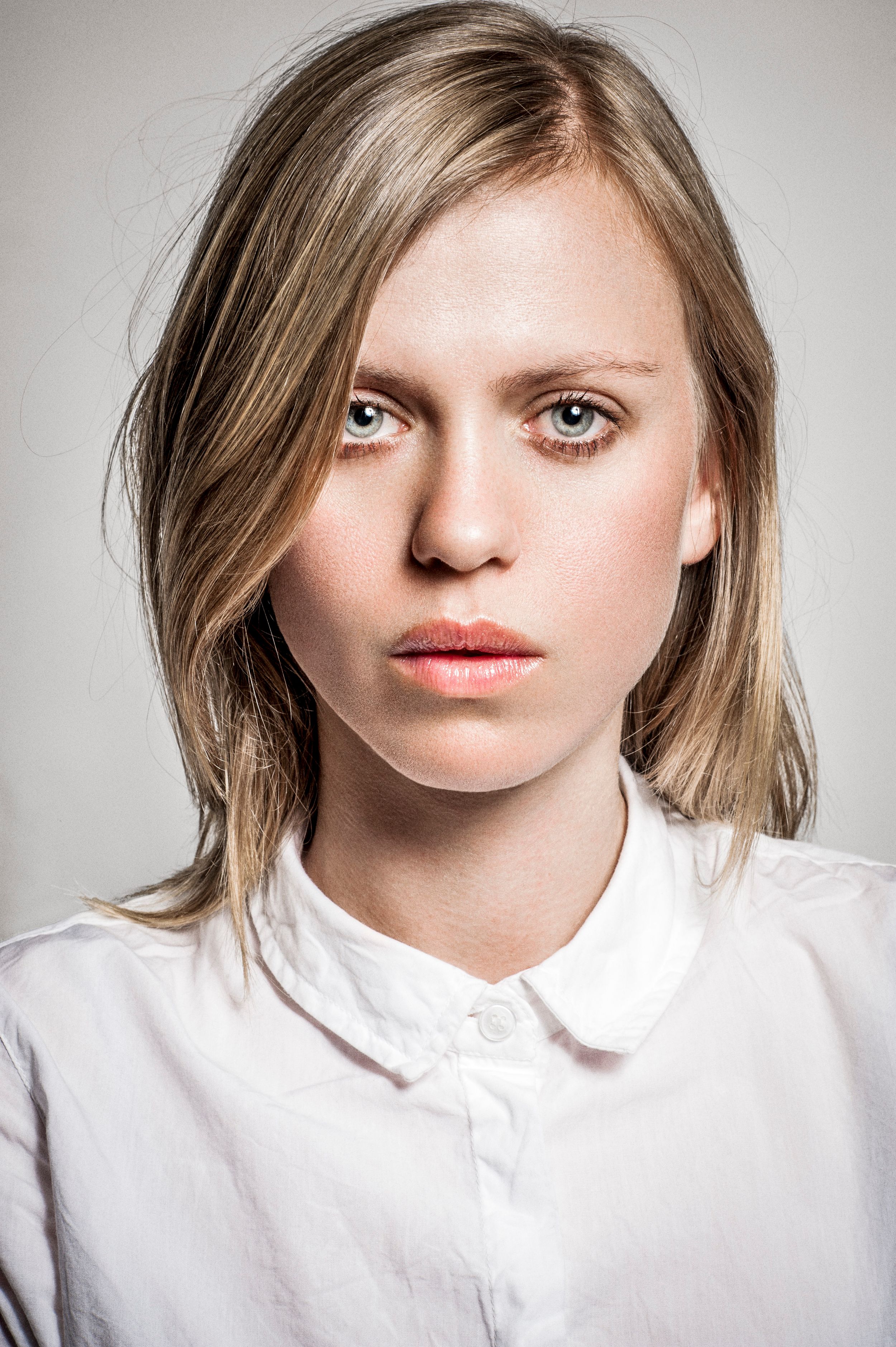 Young woman with blonde hair and blue eyes wearing a white collared shirt, neutral expression, studio portrait.