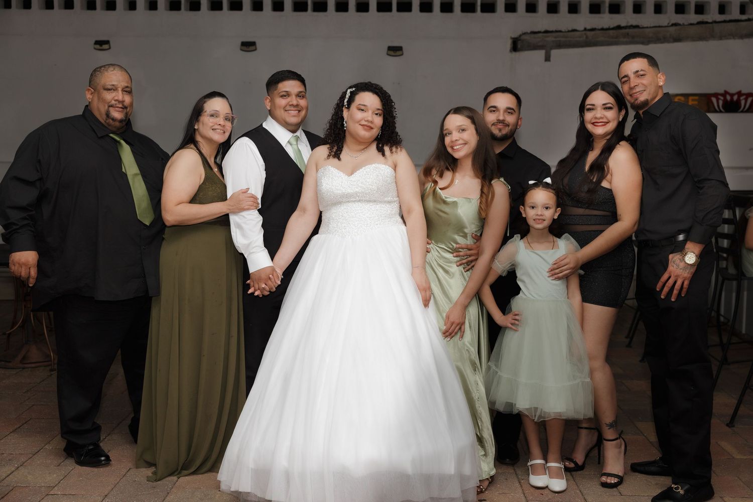 A wedding party in formal attire poses together, with olive green bridesmaid dresses and a white tulle bridal gown.