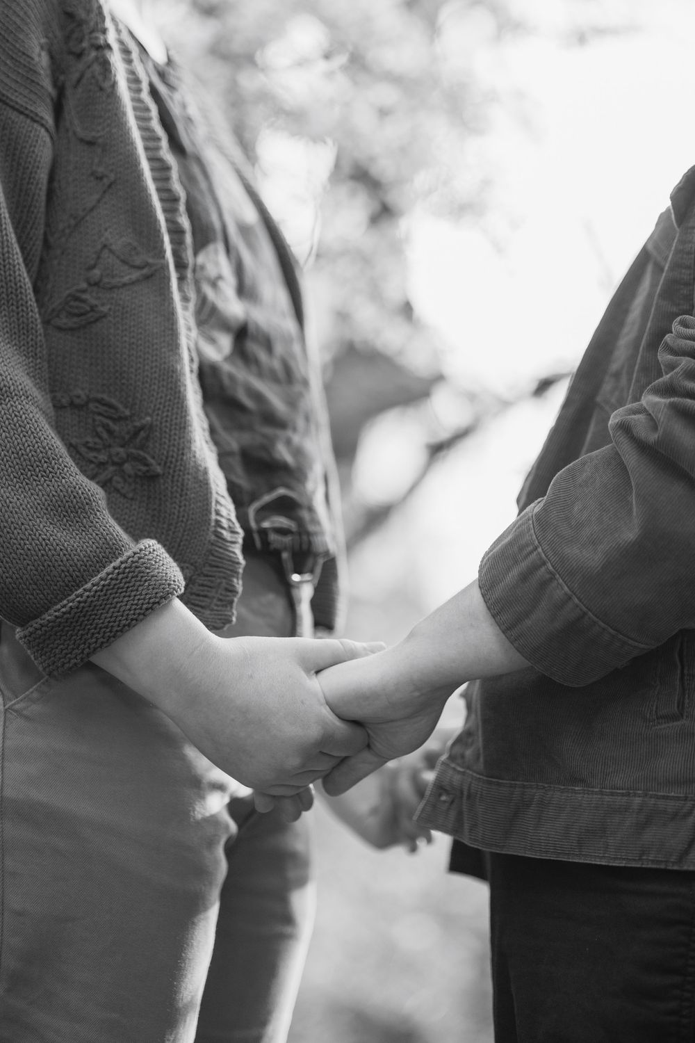 Black and white close-up of two people holding hands tenderly outdoors among blurred trees.