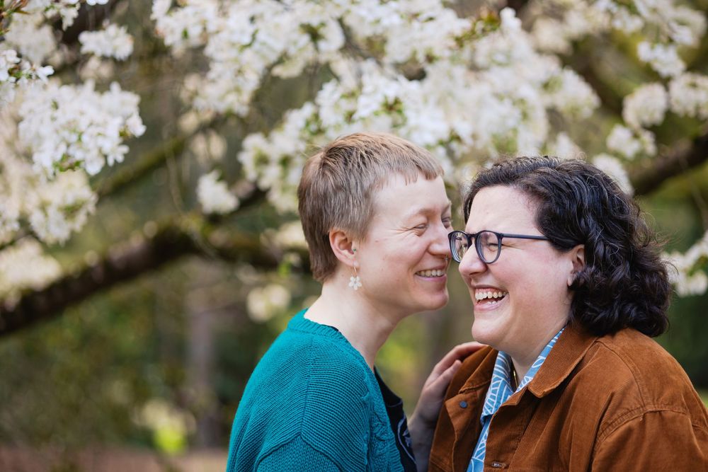 Two women smile and touch noses affectionately beneath white cherry blossoms in spring garden.