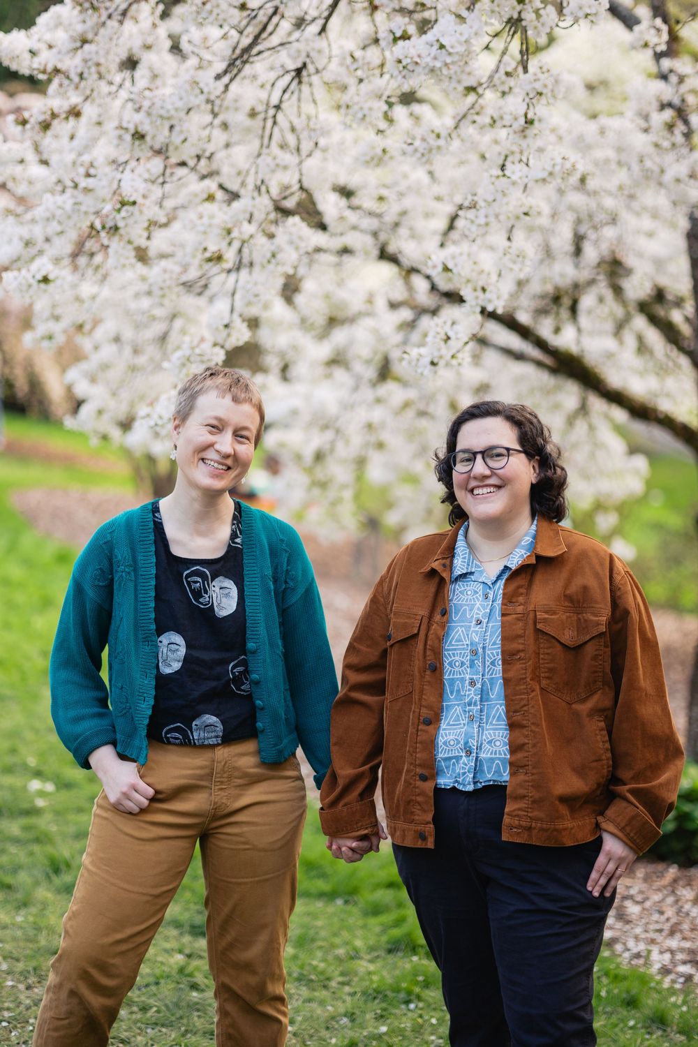 Two women hold hands and smile while standing before white cherry blossom trees in spring.
