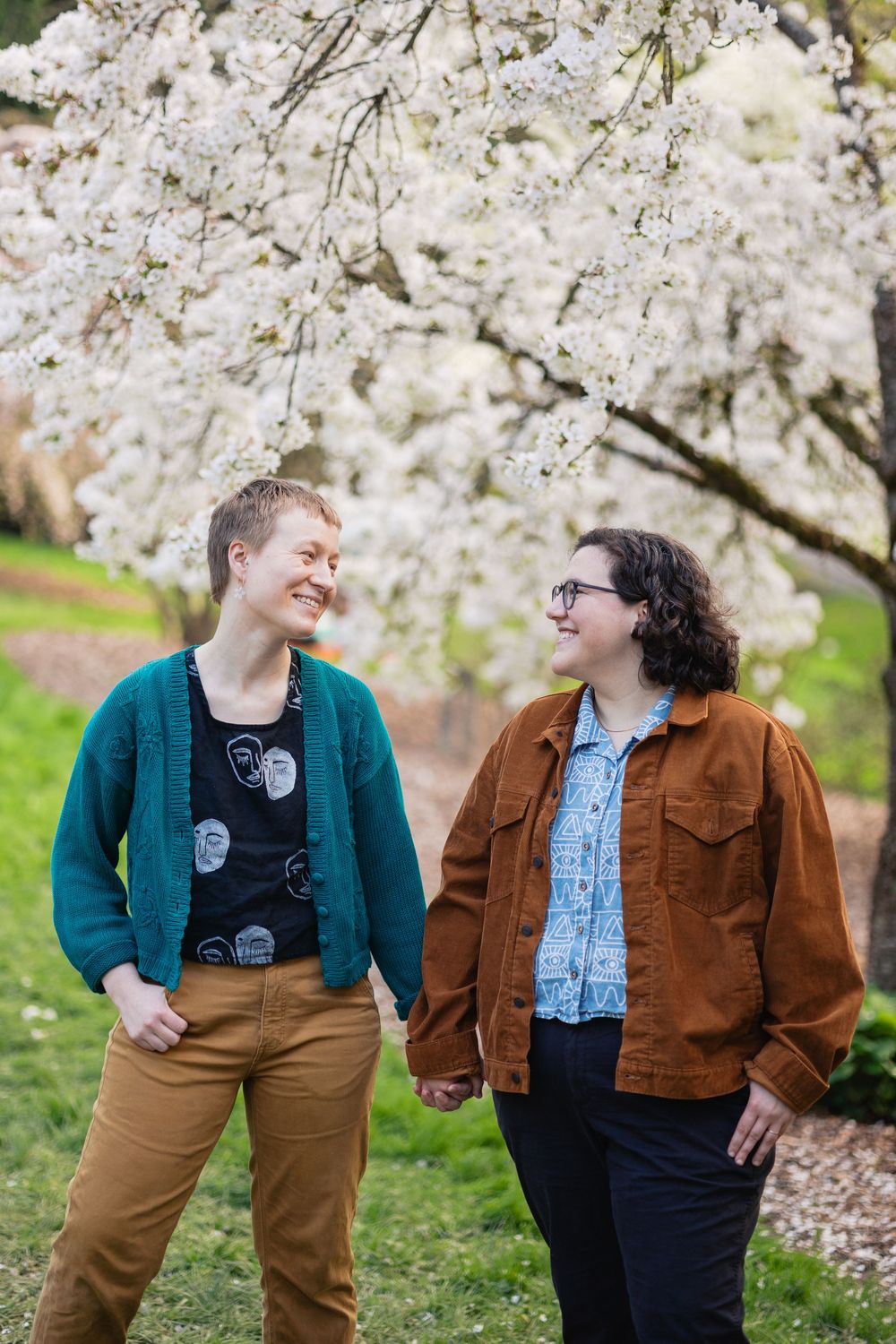 Two women hold hands and smile at each other beneath a blooming white cherry blossom tree in spring.
