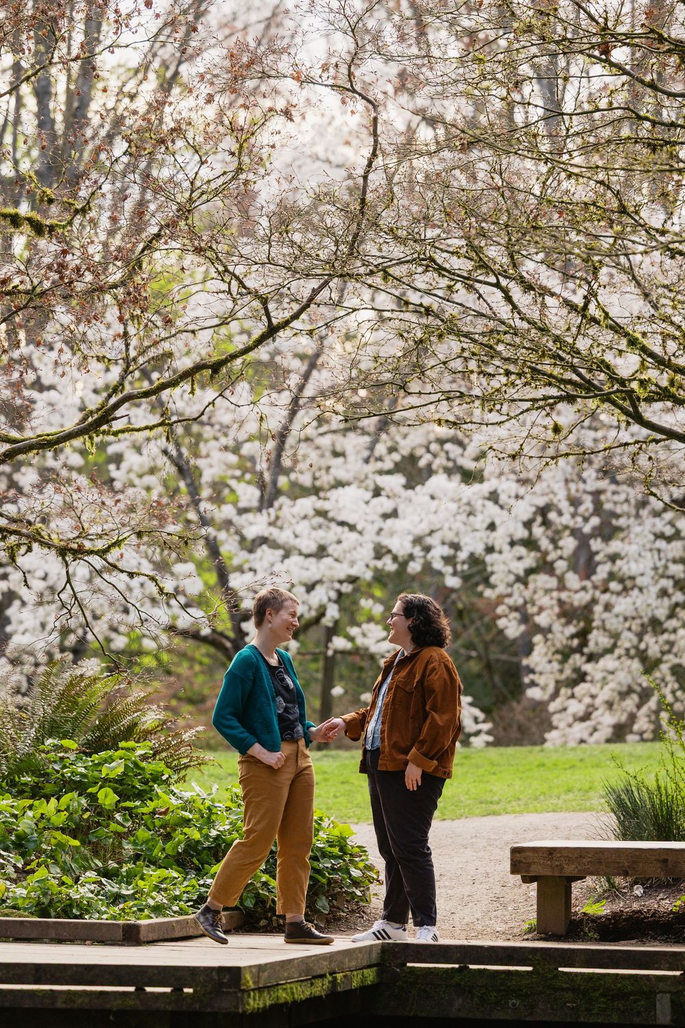 Two women share a conversation on a garden path surrounded by lush white flowering trees in spring.