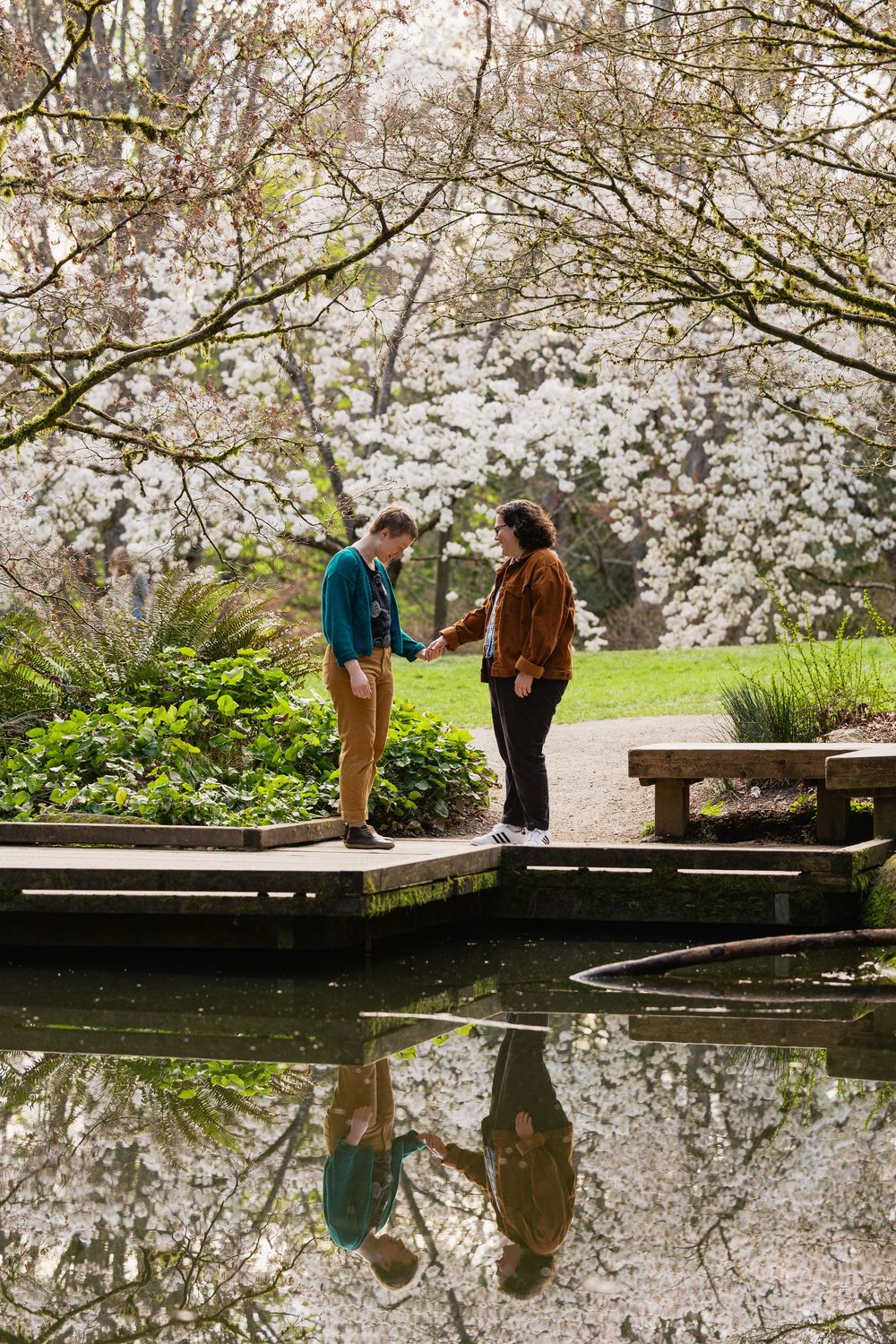 Two women hold hands on a wooden deck reflected in a still pond beneath white blossoming trees.