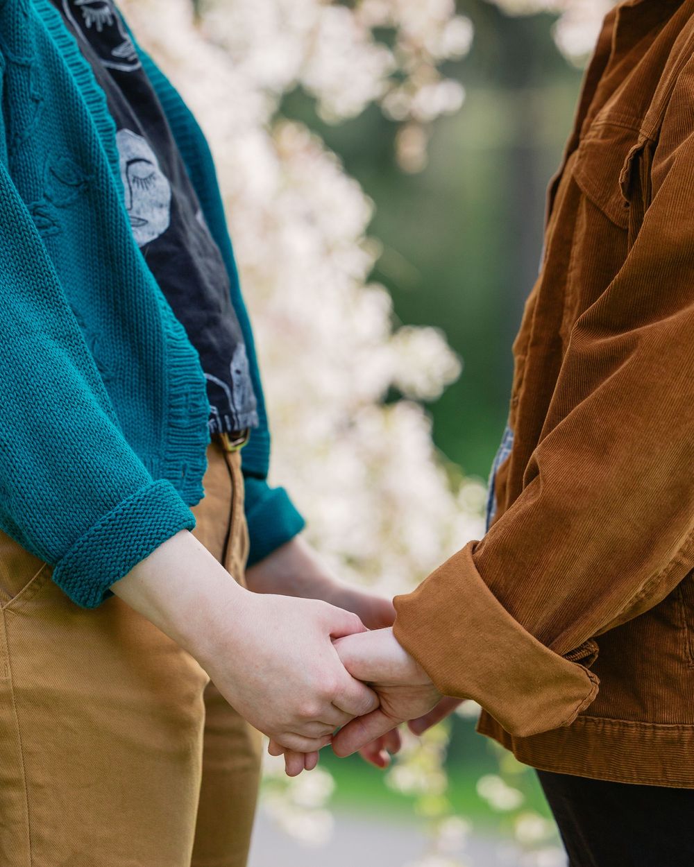 Close-up of two women holding hands, wearing teal and brown jackets with blossoms in background.
