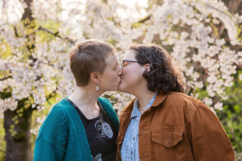 Two women share a romantic kiss in front of stunning white cherry blossom trees in spring.