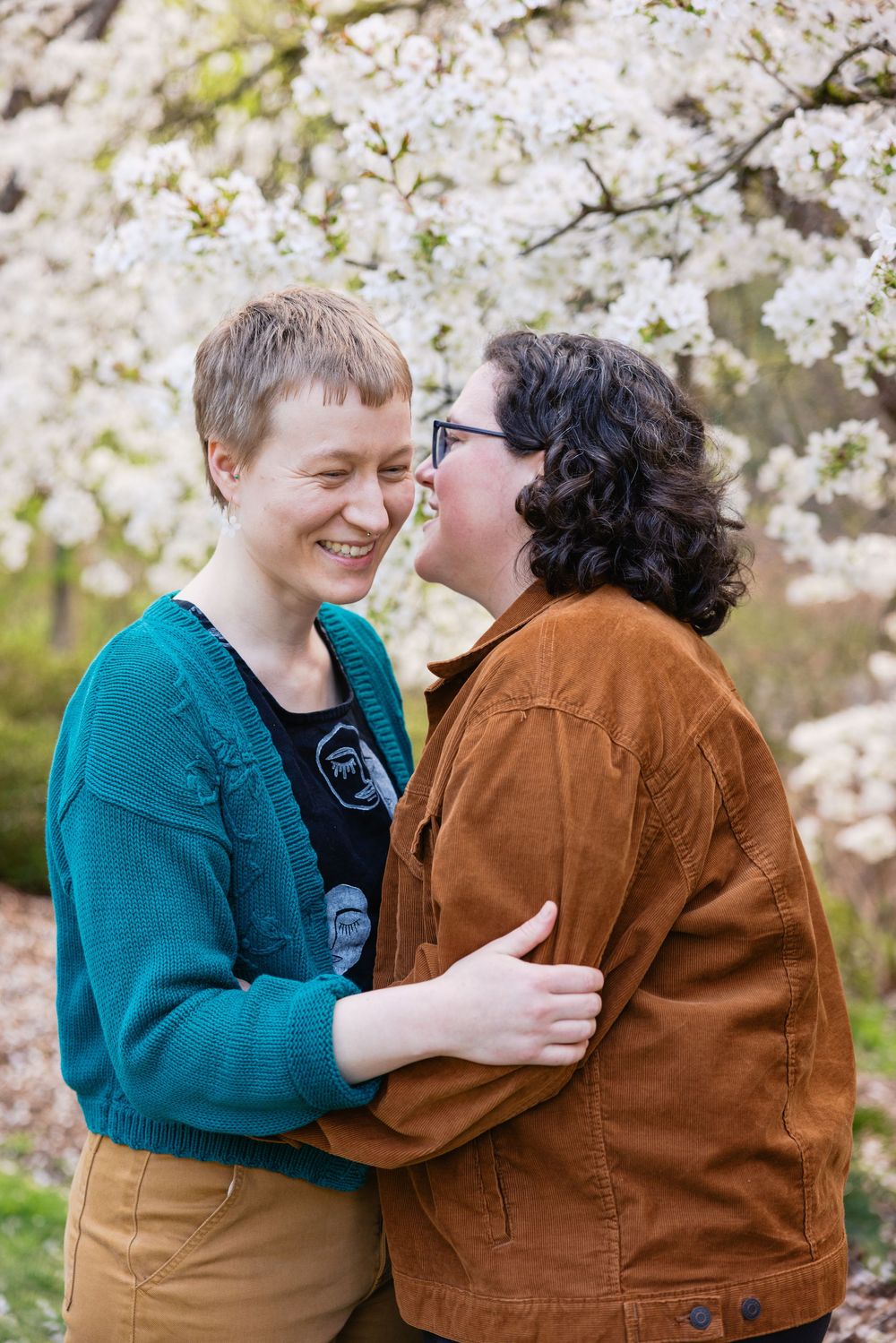 Two women embrace and laugh joyfully together under blooming white cherry blossom trees outdoors.