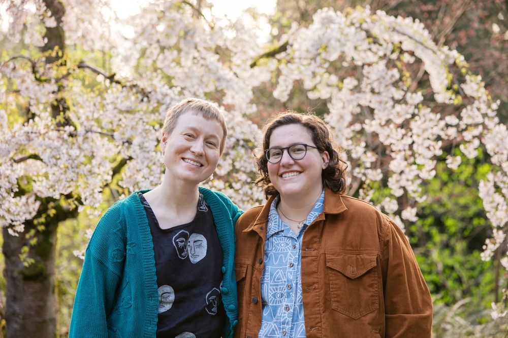 Two smiling women stand together in front of beautiful white cherry blossom trees in spring.