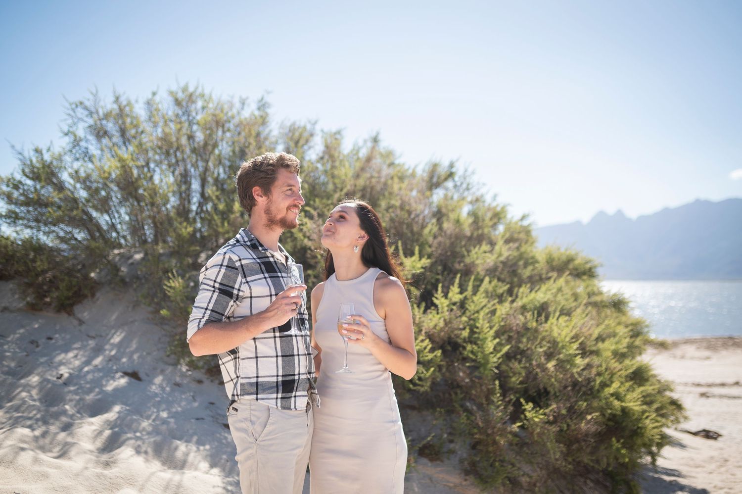 Couple smiling at each other on sandy beach with mountains and blue sky backdrop.